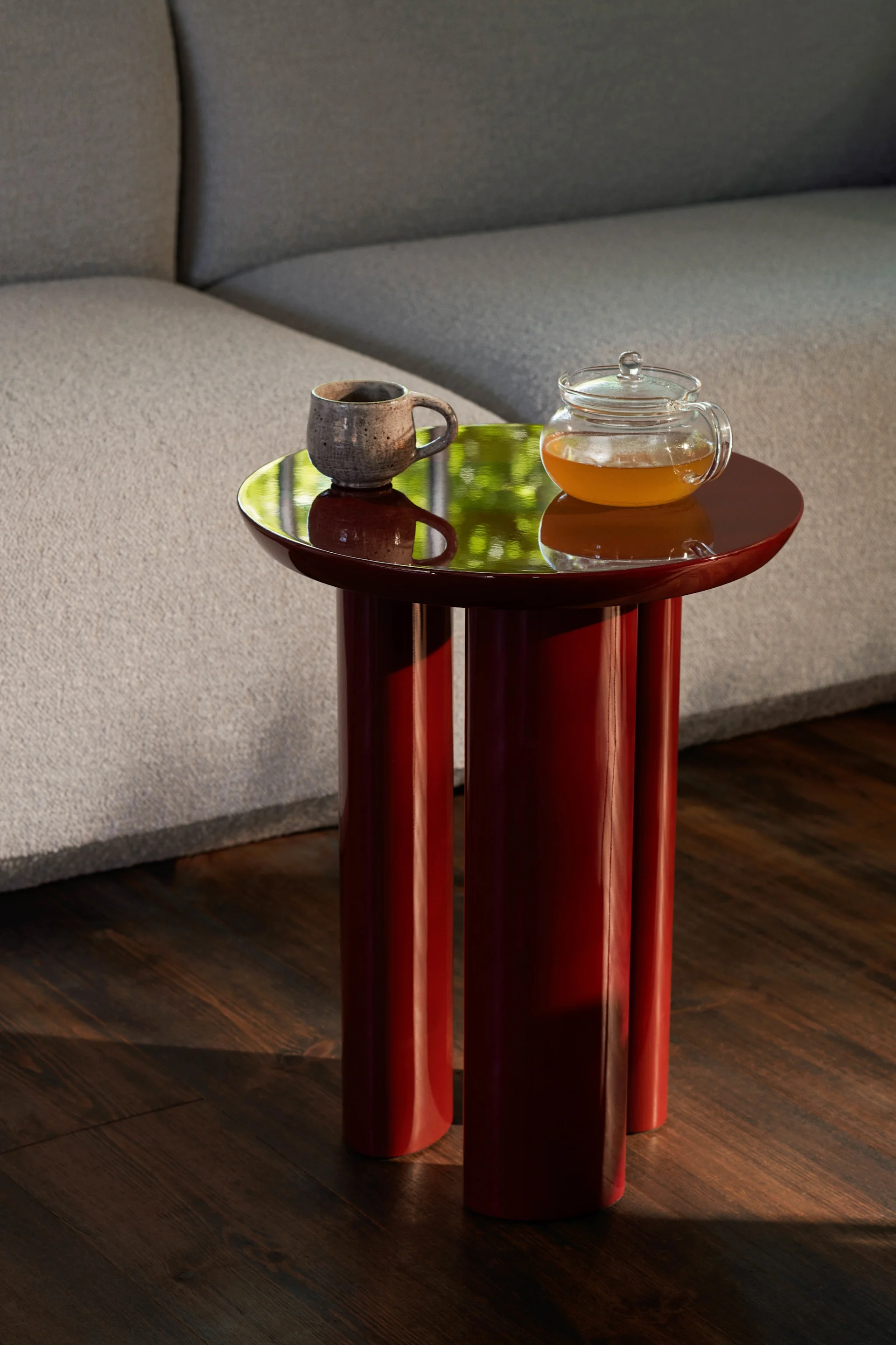 Mesa auxiliar roja brillante con tres patas, con una taza de cerámica y una tetera de cristal llena de té ámbar.