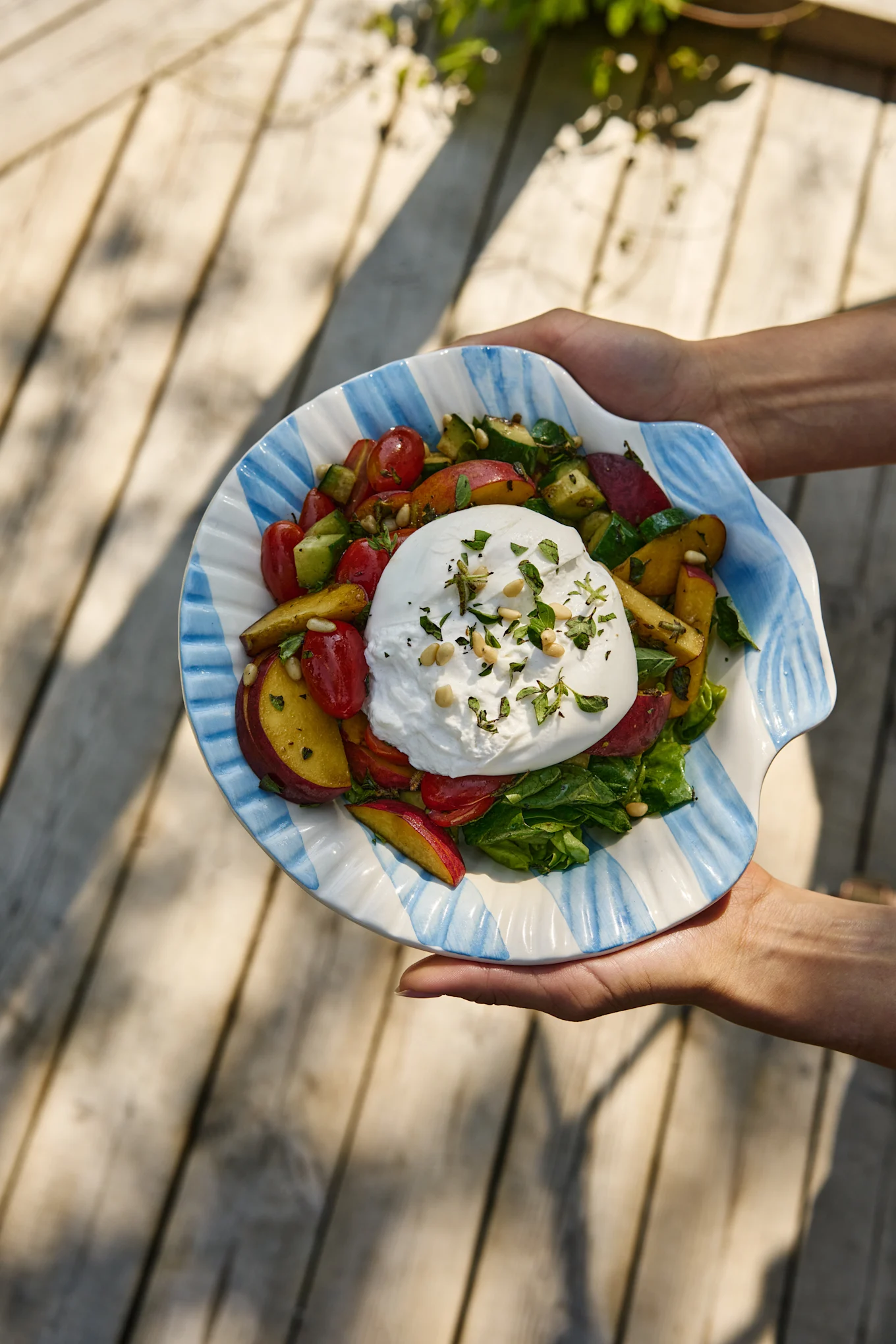 Manos sostienen una ensalada de burrata con tomates, duraznos, pepino y verduras en un cuenco rayado con forma de concha.