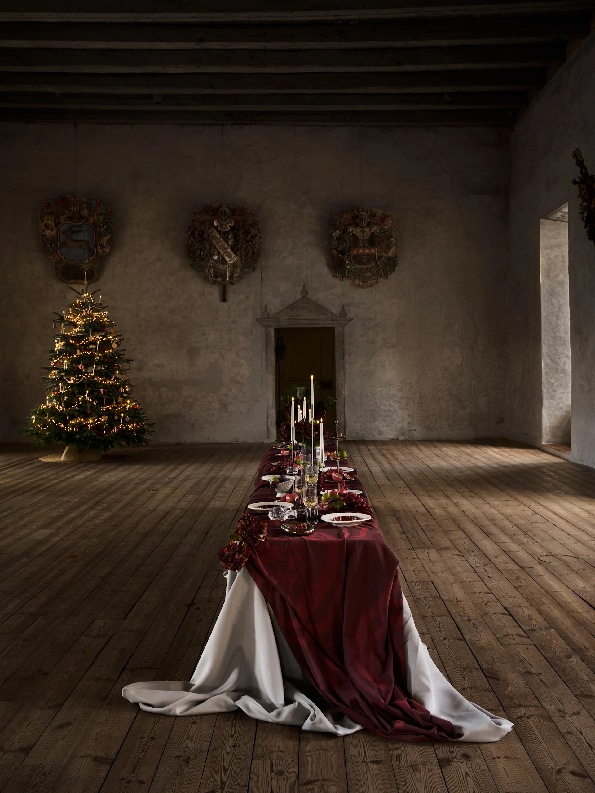 Larga mesa de cena navideña con mantel rojo, velas, fruta y cubiertos, junto a un árbol de Navidad iluminado en un salón antiguo.