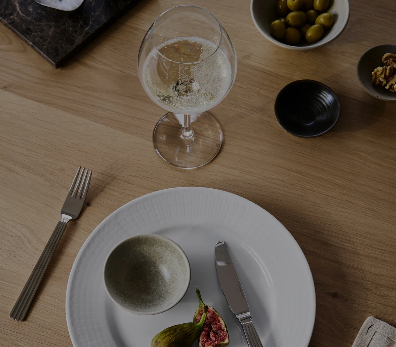 Una copa de champán, plato blanco con un cuenco y higos, tenedor, cuchillo, aceitunas y nueces en una mesa de madera.