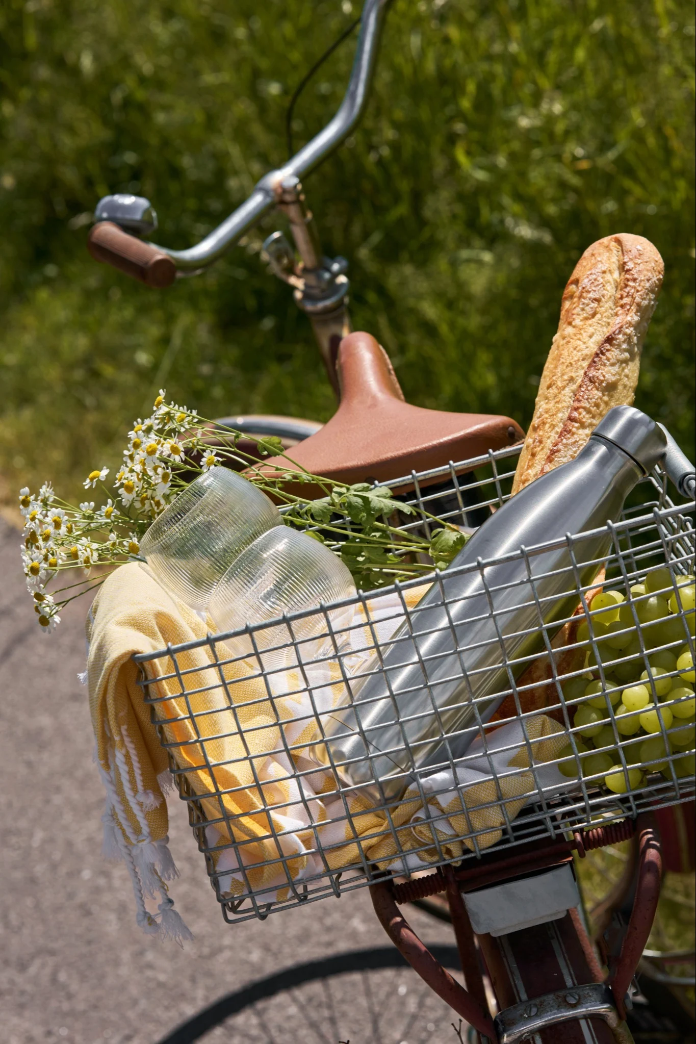 Bicicleta con una cesta delantera llena de artículos de picnic: pan, uvas verdes, una botella de agua plateada, vasos, una toalla amarilla y flores.