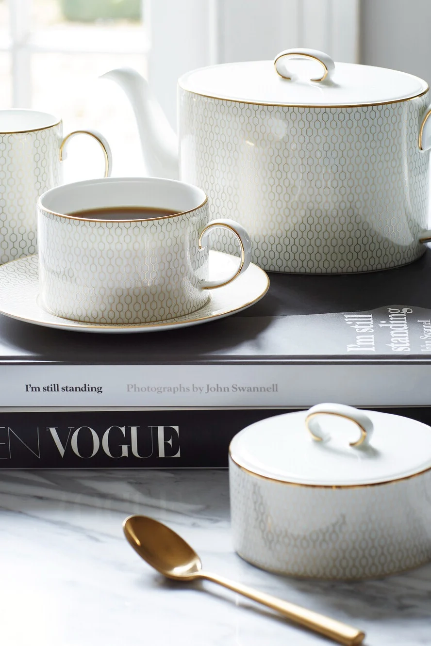 Juego de té de porcelana blanca con patrón geométrico dorado sobre libros, que incluye tetera, taza de té, azucarero y una cuchara dorada.