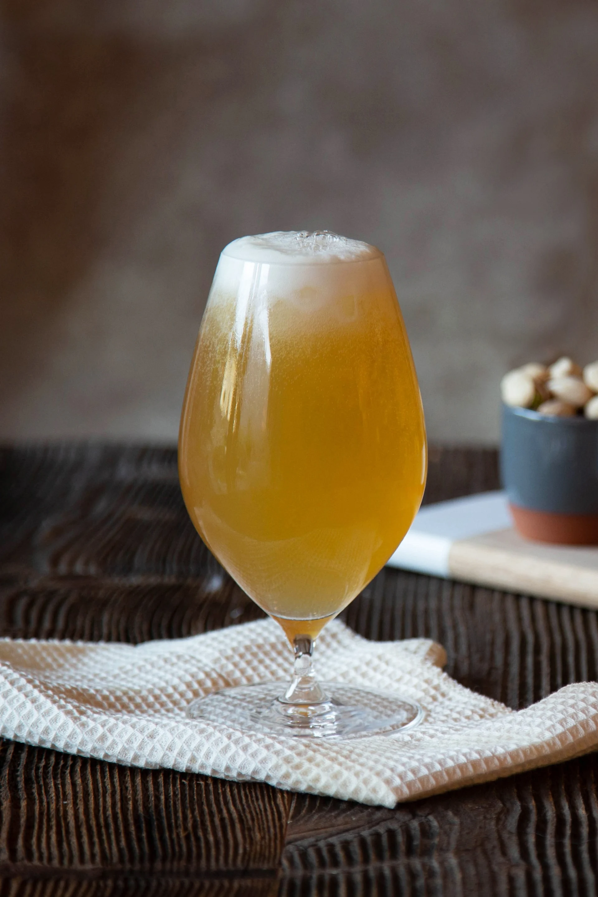 Un vaso de cerveza ámbar espumosa sobre un paño blanco, con cuencos de frutos secos y palitos de pan en el fondo sobre una tabla de madera.