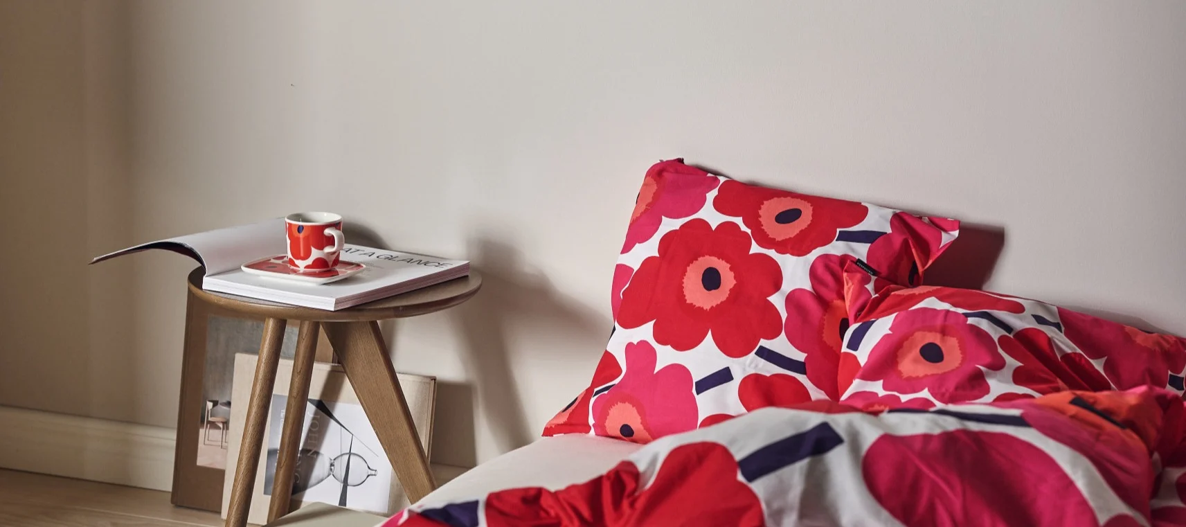 Cama con ropa de cama floral roja, una taza a juego en una mesa auxiliar con una revista.