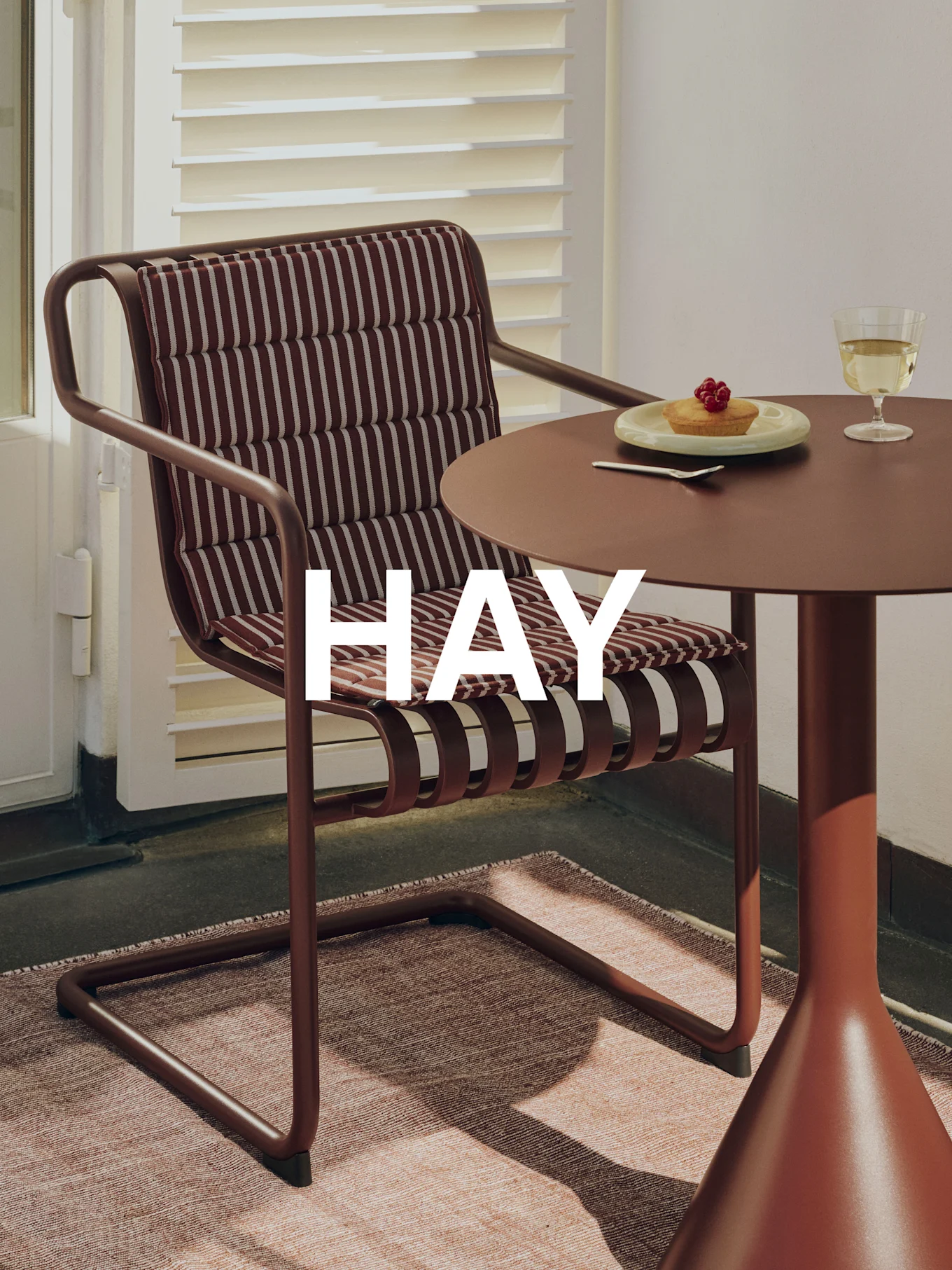 Ambiente exterior con silla de metal marrón, cojín a rayas, mesa a juego, tarta, copa de vino y marca 'HAY'.