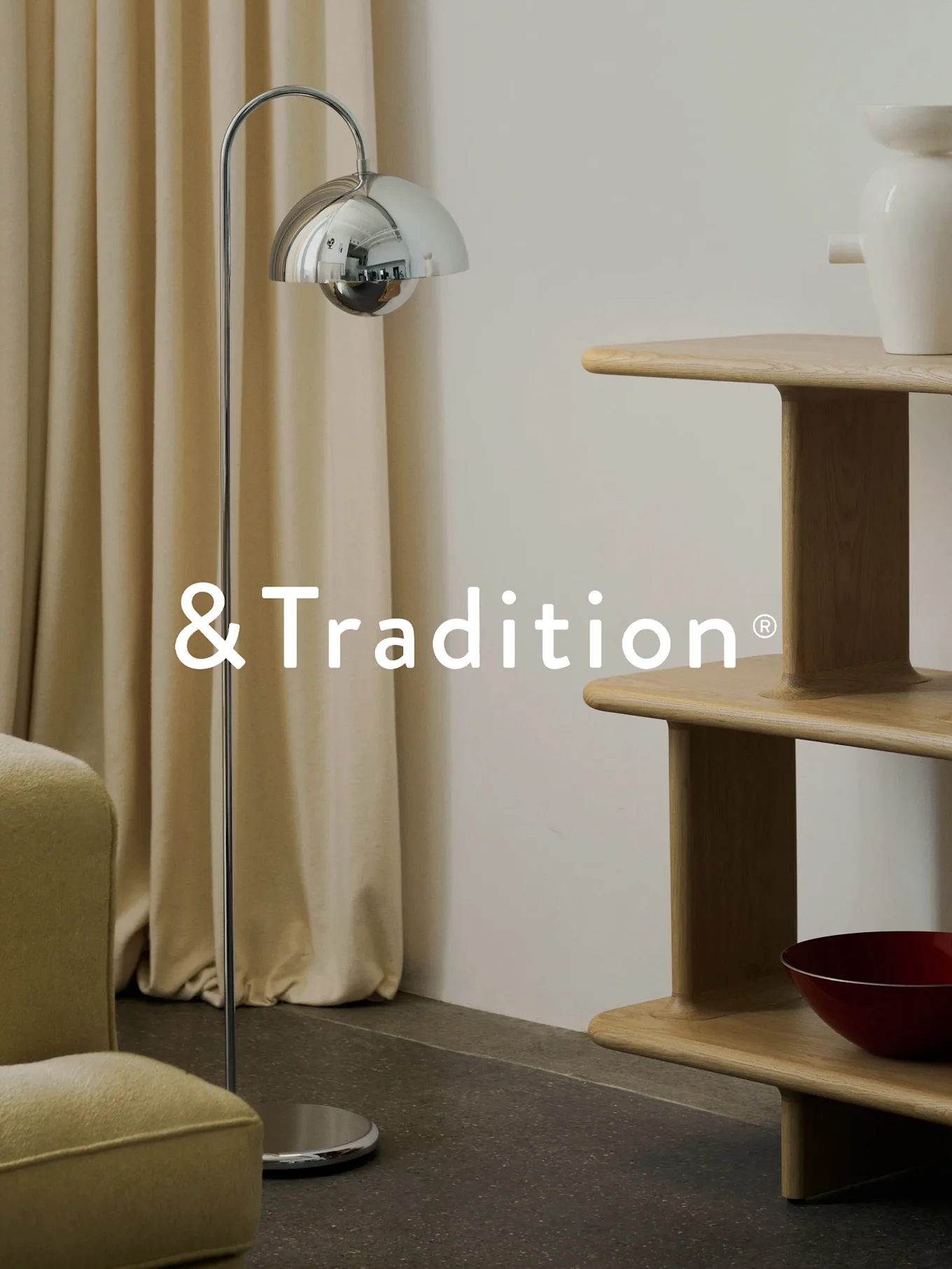 Un interior moderno con una lámpara de pie cromada, una estantería de madera clara de varios niveles y un sillón. Logotipo de &Tradition®.