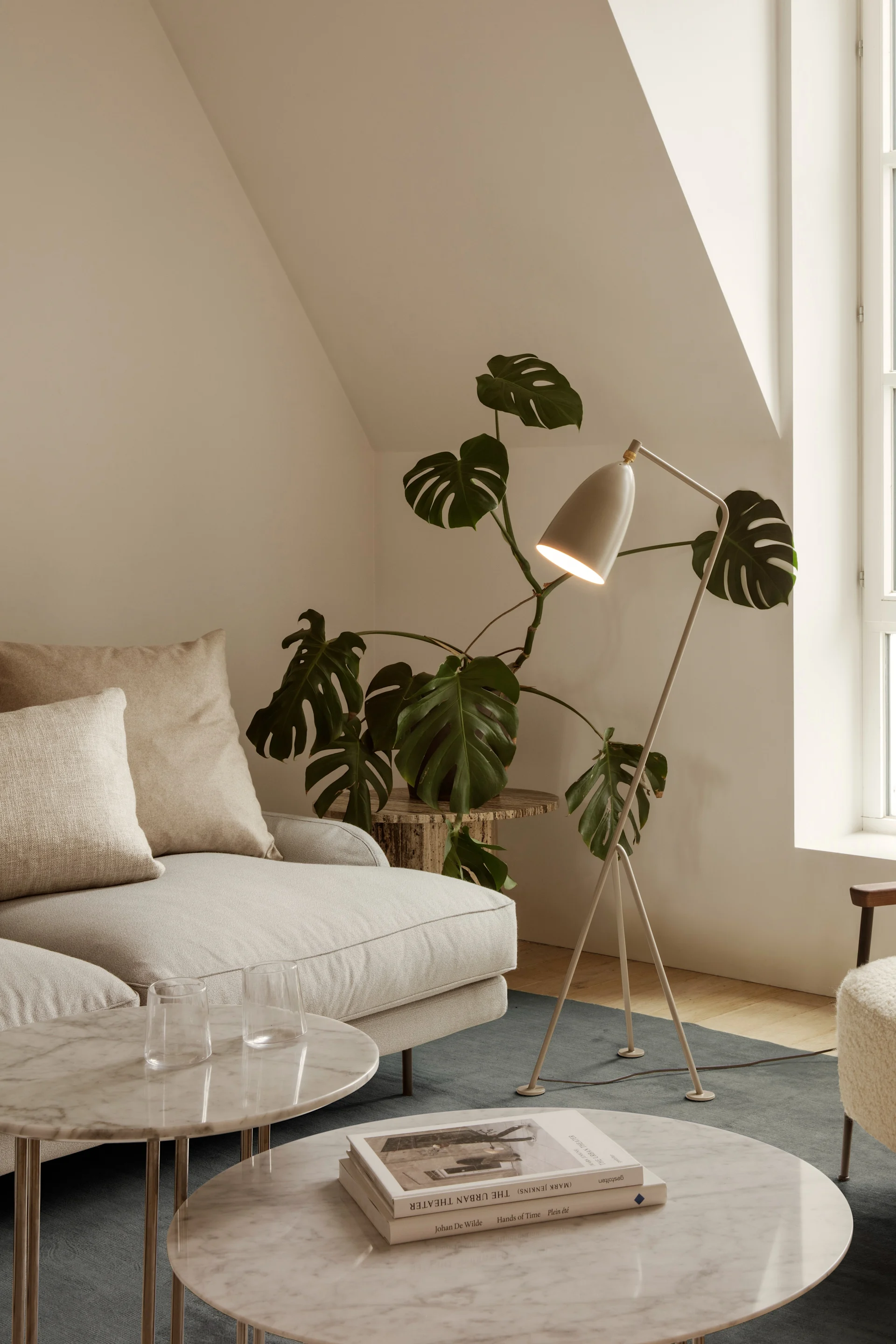 Sala de estar luminosa con sofá crema, mesas mármol, lámpara de pie, planta y sillón mullido junto a ventana arqueada.