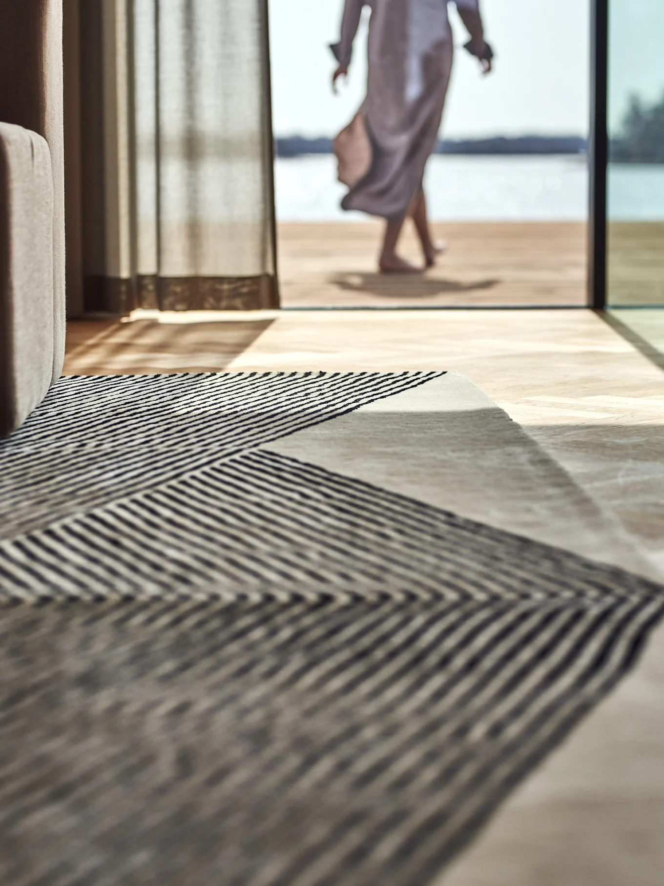 Alfombra de rayas geométricas en suelo de madera, con una persona caminando hacia un lago exterior.