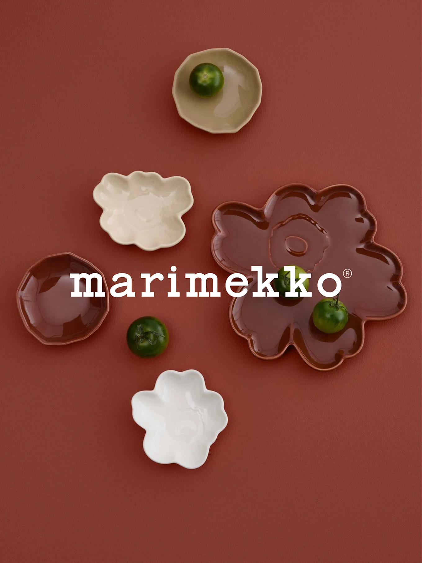Logotipo blanco 'marimekko', platos en forma de flor (blanco, crema, marrón rojizo) con frutas verdes, sobre superficie de color óxido.