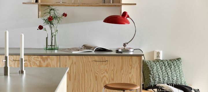 Aquí puedes ver la lámpara de mesa Kaiser en rojo rubí con el jarrón Ikeru en acero cepillado, ambos de Fritz Hansen, en una moderna cocina de madera contrachapada.
