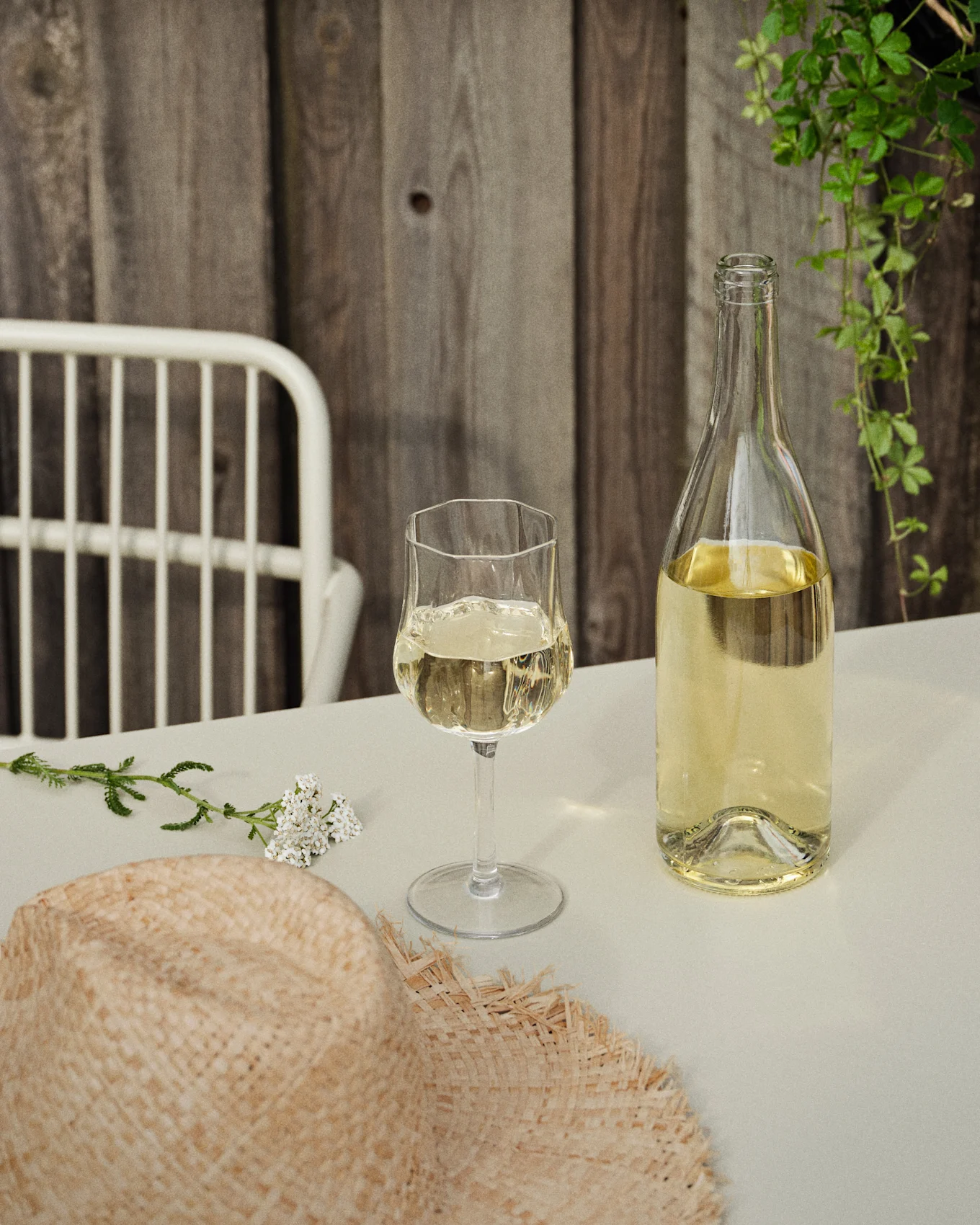Una botella y una copa de vino blanco sobre una mesa con un sombrero de paja y flores blancas, contra una pared de madera y enredaderas verdes.