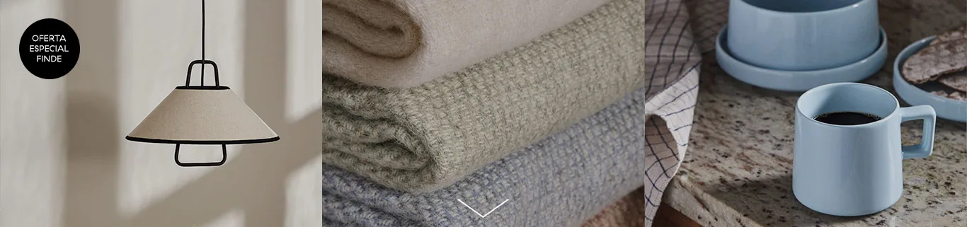 Lámpara colgante con pantalla beige, pila de mantas en beige, verde y azul, y taza de café azul en una encimera.