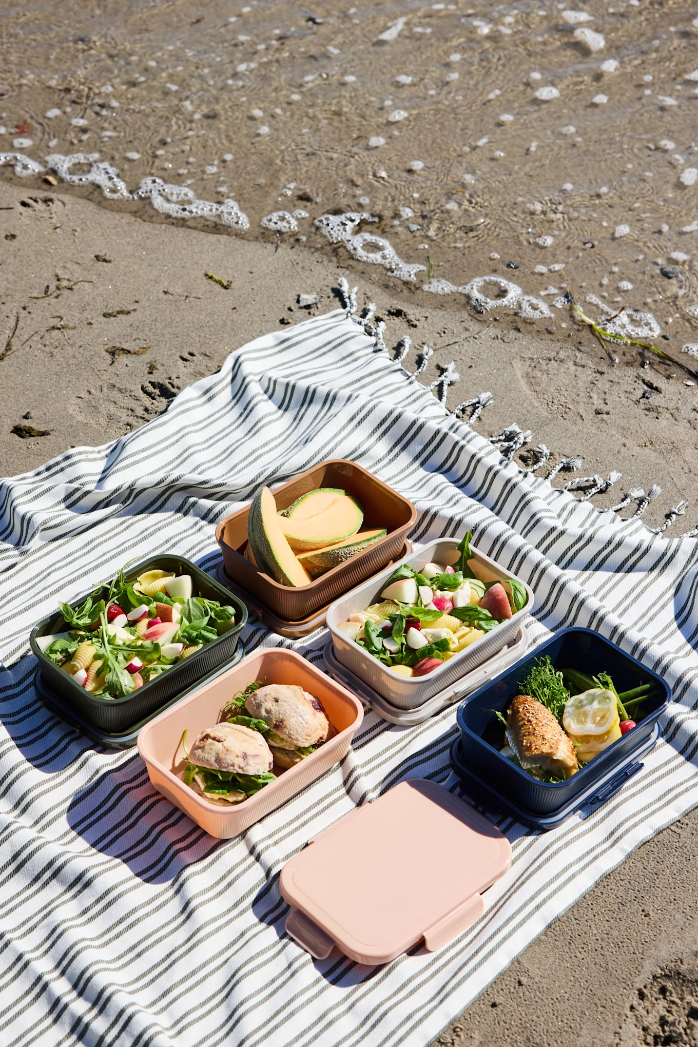 Cinco fiambreras de colores abiertas con ensaladas, sándwiches y melón, y una caja rosa cerrada, sobre una manta de rayas en la playa.