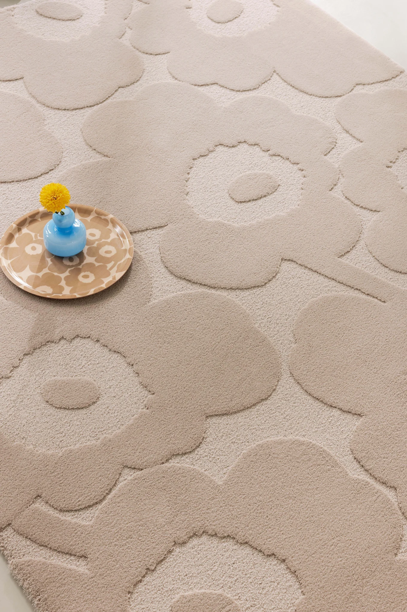 Alfombra beige con un motivo floral en relieve, en la que se ve un jarrón azul claro con una flor amarilla sobre una bandeja estampada.