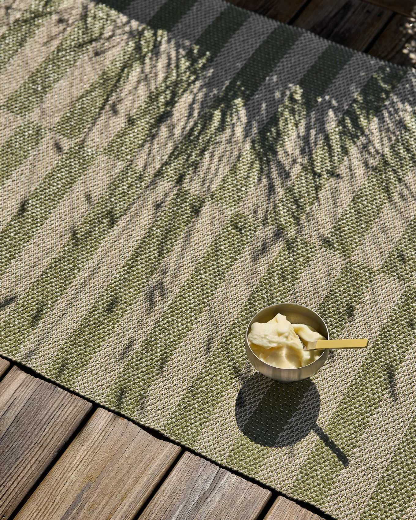 Alfombra tejida a rayas verdes y beige con un tazón de helado y sombras de palma en una terraza de madera.