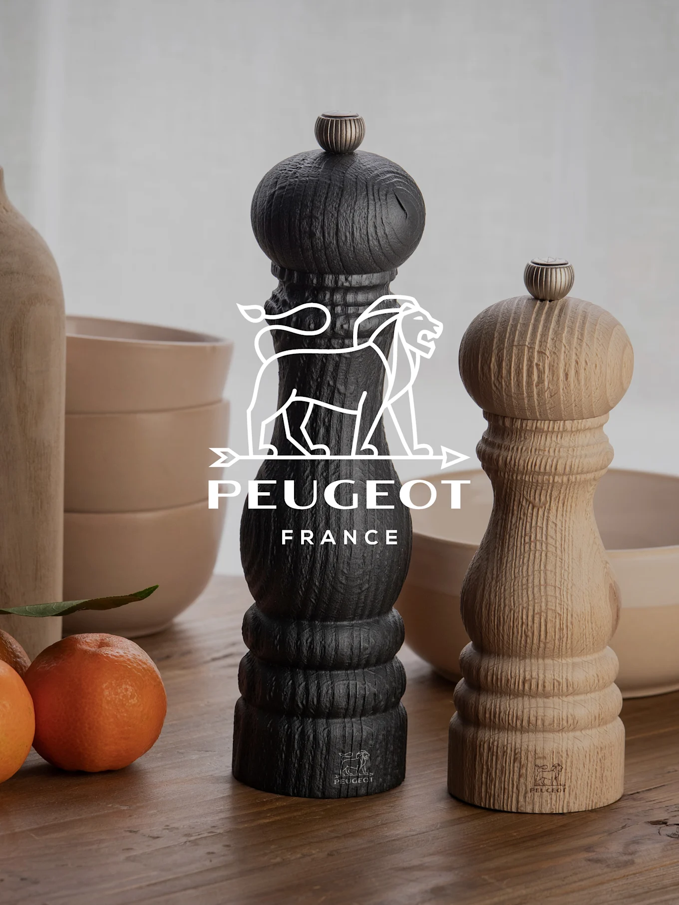 Dos molinillos de pimienta Peugeot, uno negro y otro de madera natural, sobre una mesa de madera con mandarinas y cuencos.