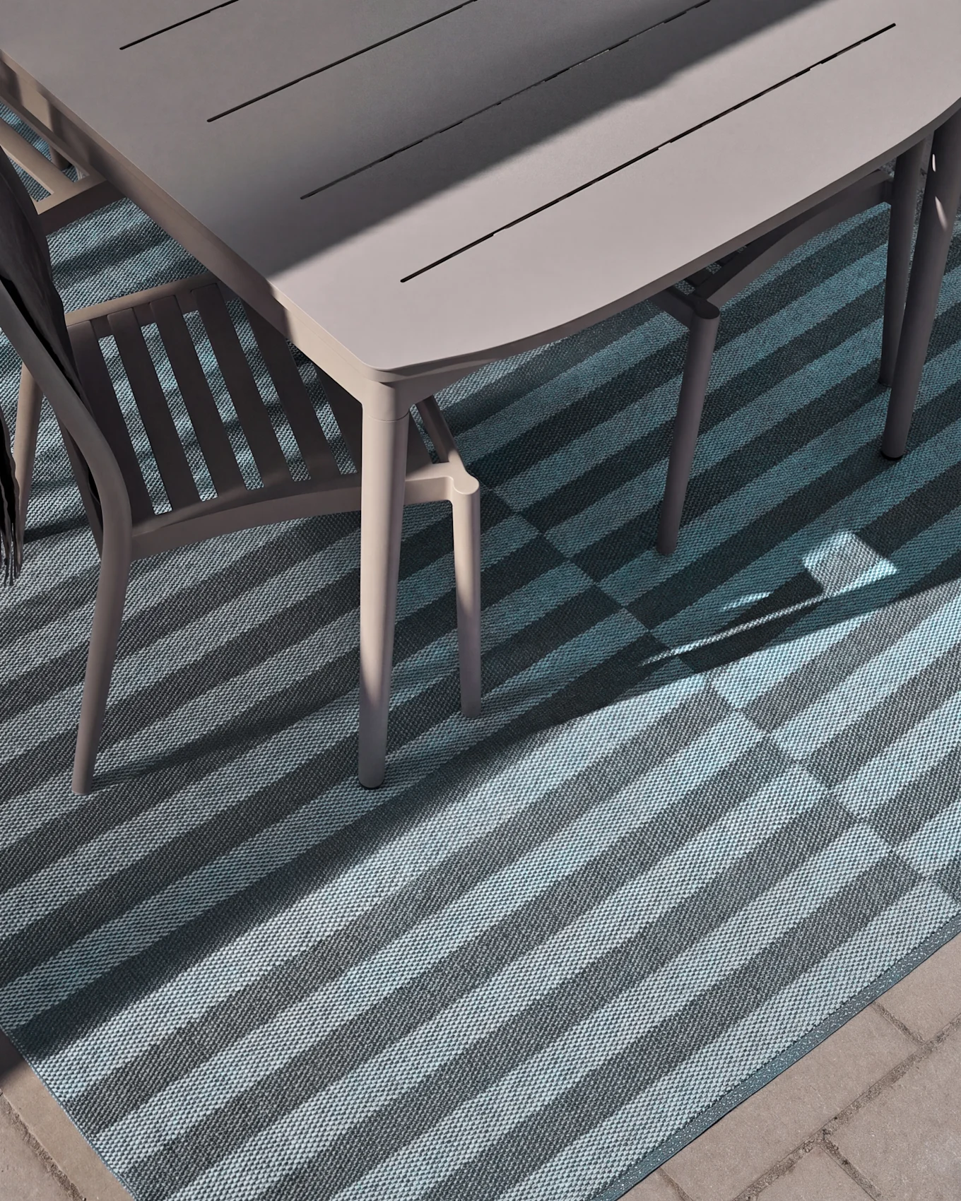 Vista superior de una mesa y silla de comedor exterior sobre una alfombra a rayas con franjas azules claras y oscuras, parcialmente iluminadas por el sol.