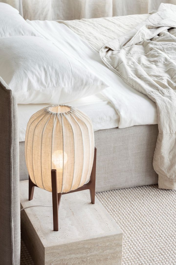 Los muebles y accesorios fabricados con materiales naturales como la tela y la madera son perfectos para un estilo inspirado en Lagom. Aquí puedes ver la lámpara de mesa Prisma de Watt & Veke junto a una cama.