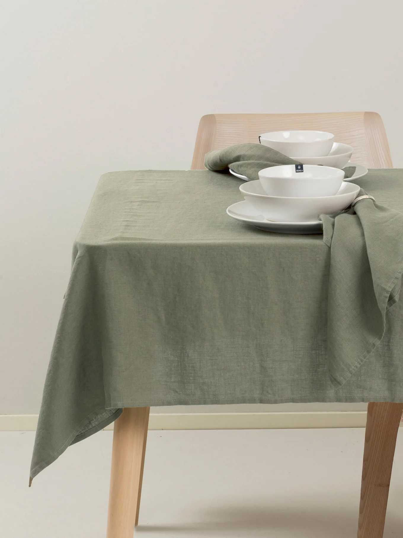 Mesa puesta con mantel y servilletas de lino verde, platos y cuencos de cerámica blanca y una silla de madera.