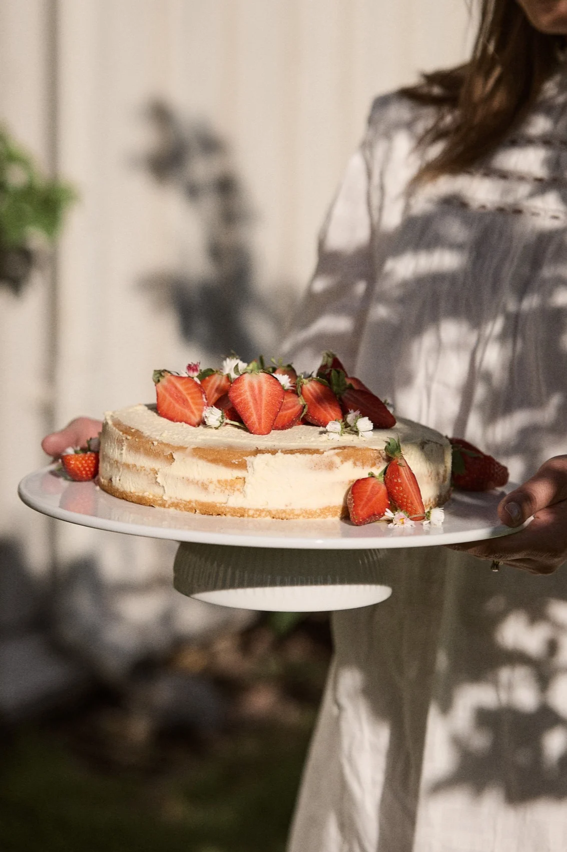Persona sosteniendo un pastel redondo cubierto con fresas frescas partidas por la mitad y pequeñas flores blancas en un soporte para pasteles blanco al aire libre.