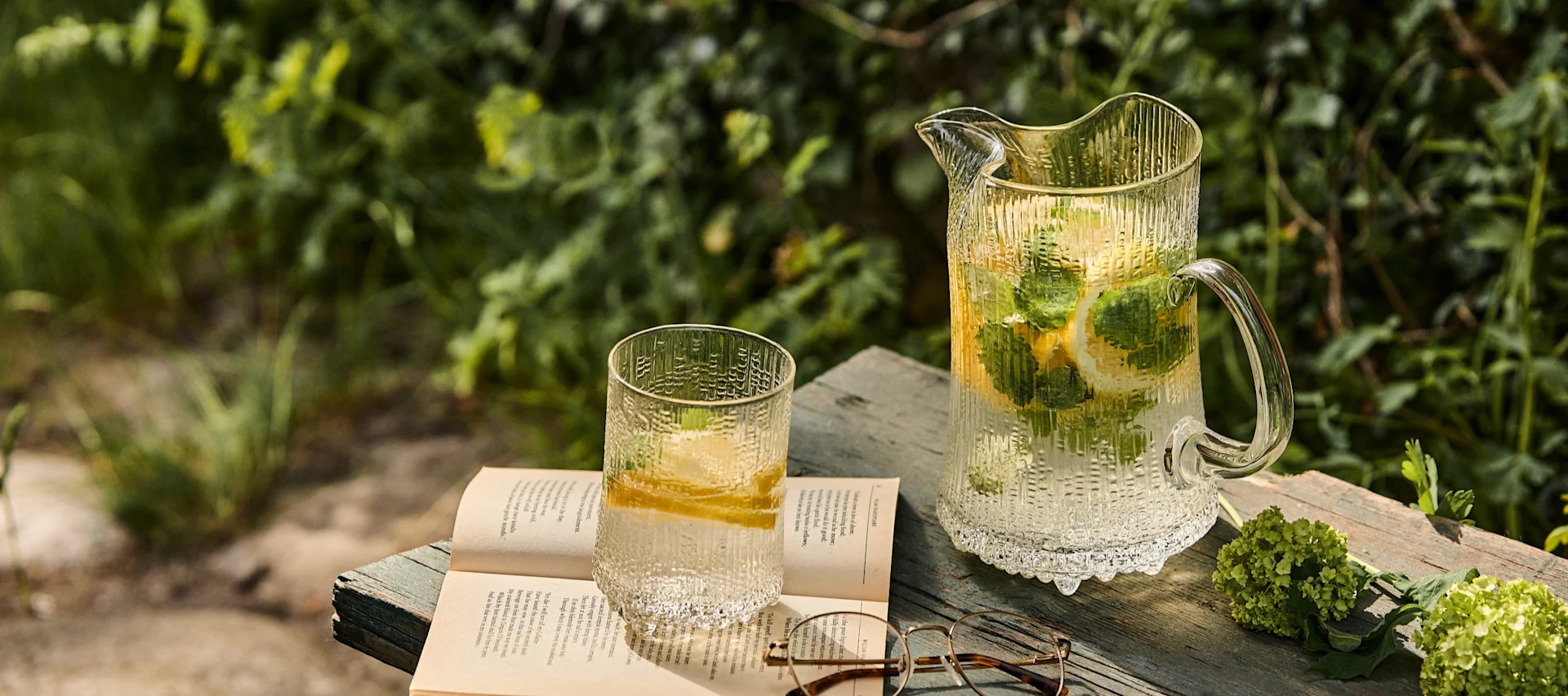 Jarra y vaso de cristal texturizado con agua de limón y menta, un libro abierto y gafas sobre una superficie de madera al aire libre.