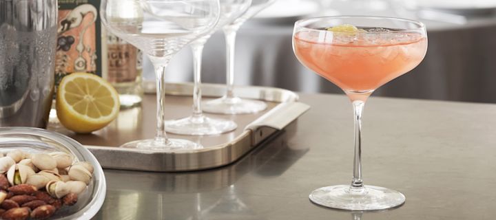 Un cóctel sin alcohol con zumo de pomelo en una copa de cóctel Cabernet de Holmegaard sobre una encimera de cocina, con utensilios de cóctel al fondo.