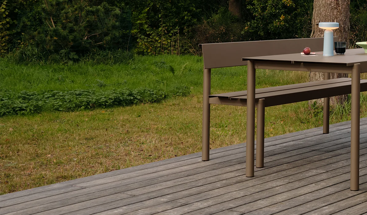 Mesa y banco de comedor modernos de color topo en una terraza de madera, con una lámpara portátil y manzanas sobre la mesa.