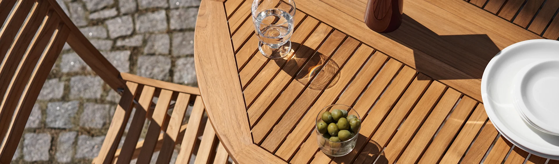Vista aérea de una mesa de patio de lamas de madera con un vaso de agua, un tazón de aceitunas verdes y platos blancos a la luz del sol.
