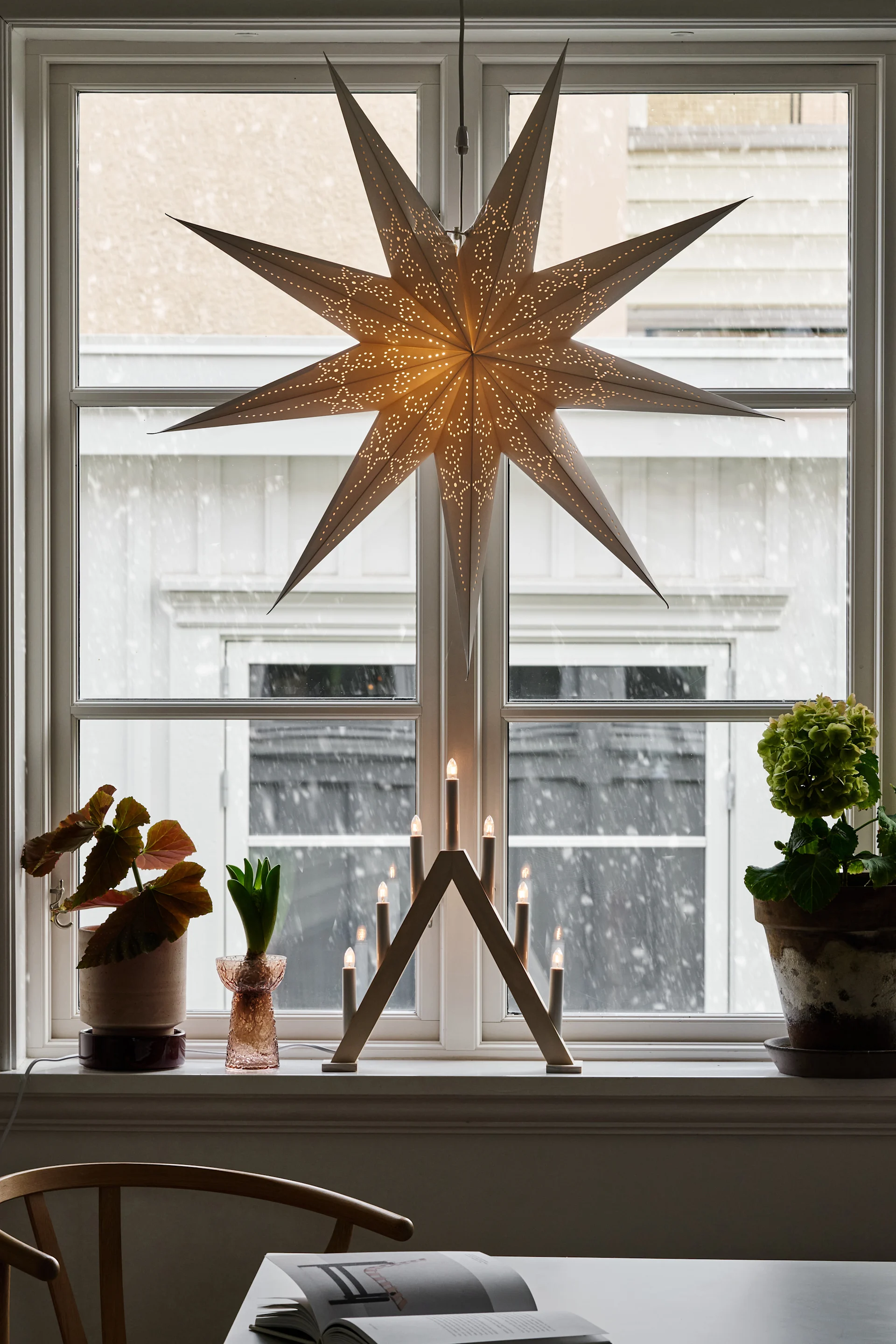 Una ventana decorada para las fiestas con una estrella de papel iluminada, un arco de madera con velas encendidas y plantas en macetas, mientras fuera cae la nieve.