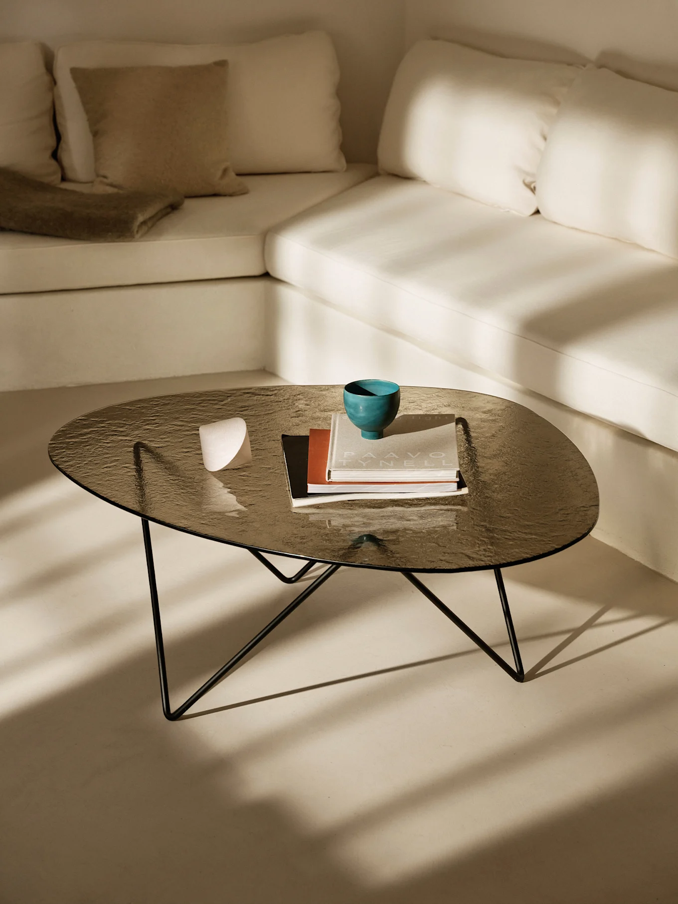 Mesa de centro de cristal oscuro con cuenco turquesa, libros y objeto blanco. Sofá en forma de L claro y sombras solares.