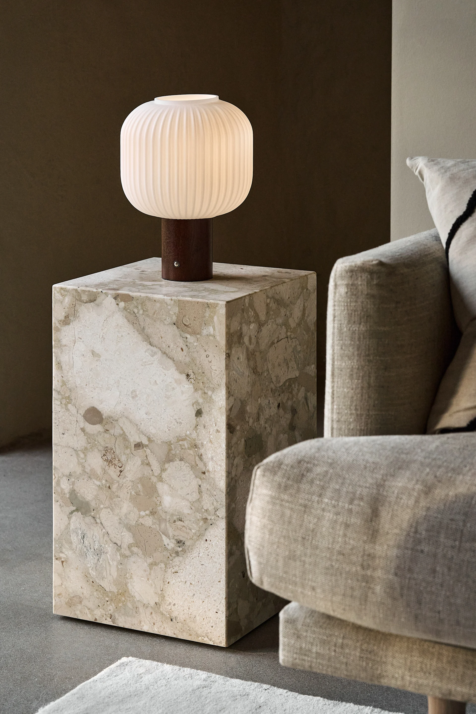 Lámpara de mesa blanca estriada y encendida con base de madera sobre una mesa auxiliar de mármol beige junto a un sofá claro.