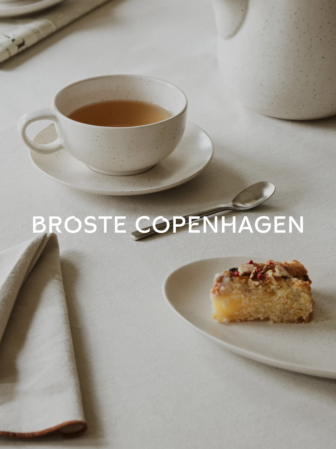 Té en taza blanca moteada, un trozo de pastel con almendras y bayas en plato a juego, cuchara y servilleta sobre mantel de lino. Texto: BROSTE COPENHAGEN.