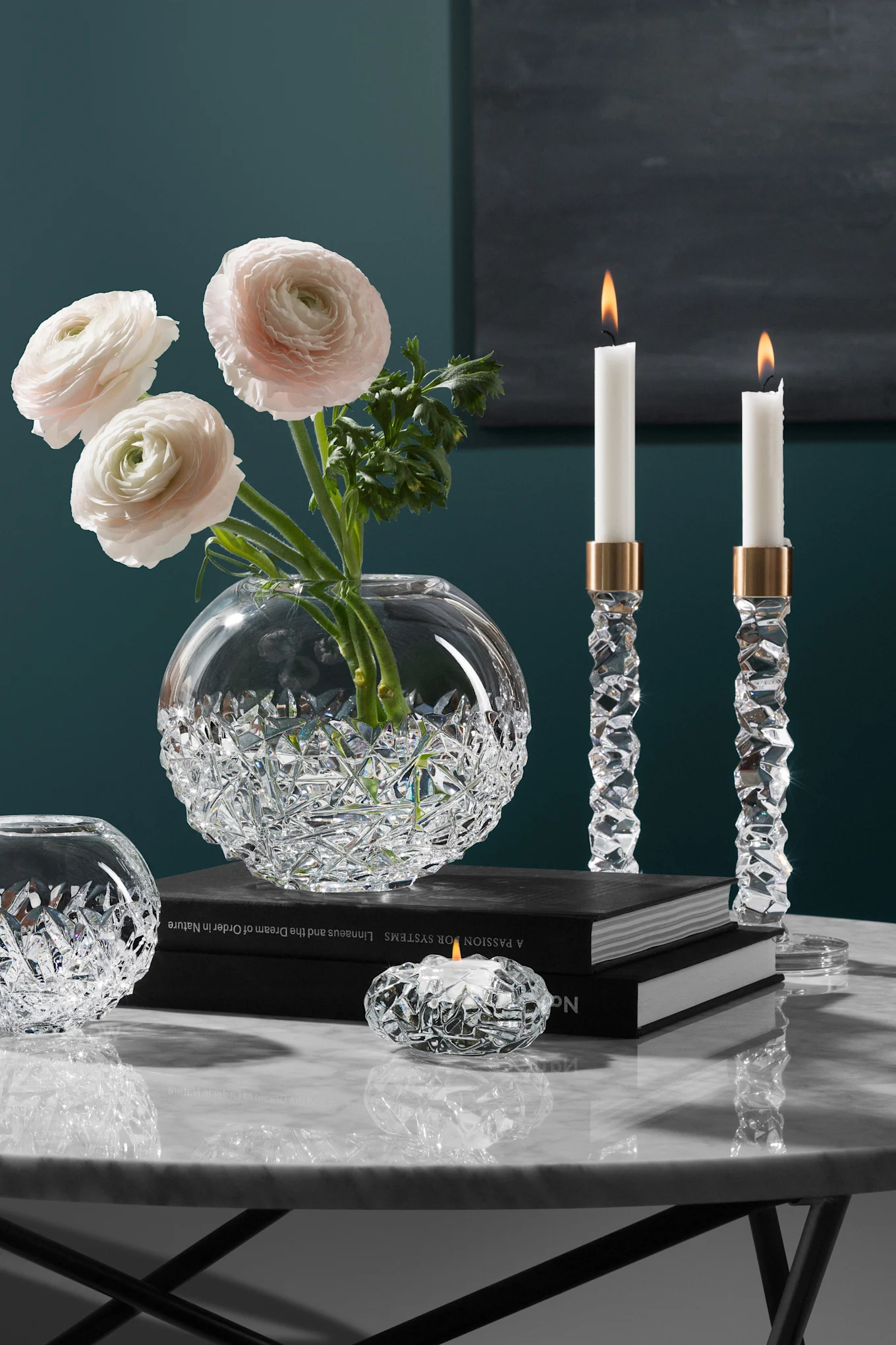 Elegante jarrón de cristal con ranúnculos rosas, dos candelabros de cristal encendidos y un portavelas en una mesa de mármol.