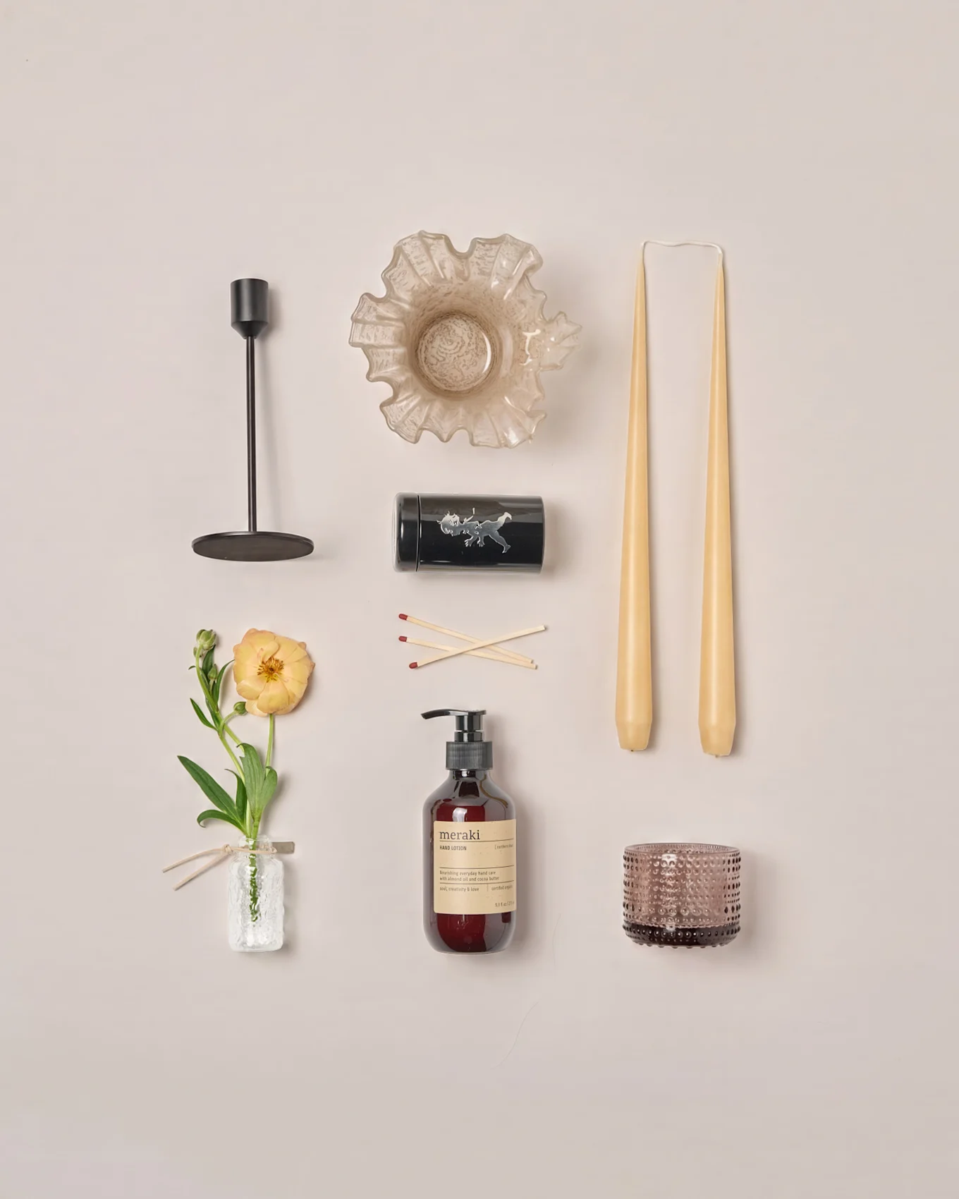 Presentación plana de varios artículos de decoración del hogar: candelabro, cuenco con volantes, velas cónicas, cerillas, loción, portavelas y una flor de ranúnculo.