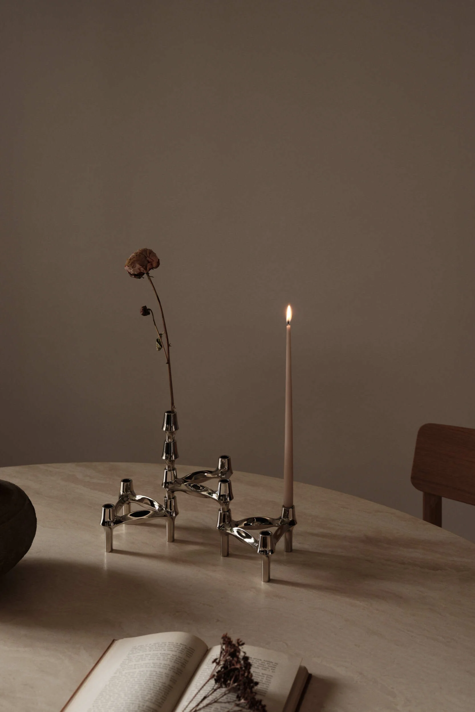 Dos candelabros modulares de tono plateado sobre una mesa redonda, uno con una vela encendida y el otro con una flor seca. Un libro abierto es visible en primer plano.