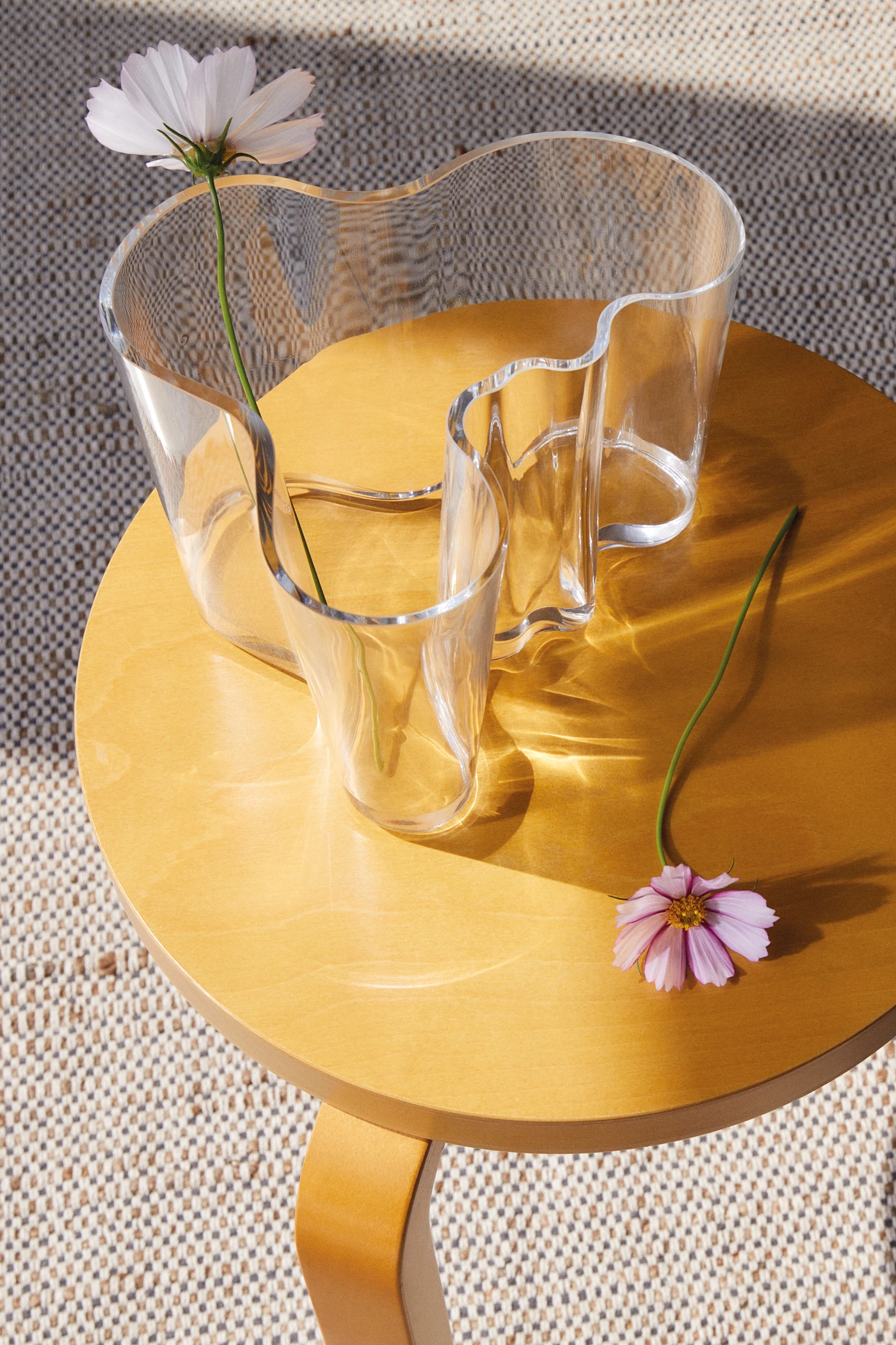 Un jarrón de vidrio ondulado y transparente con una flor blanca sobre una mesa redonda de madera, con una flor rosa al lado. El sol proyecta patrones.