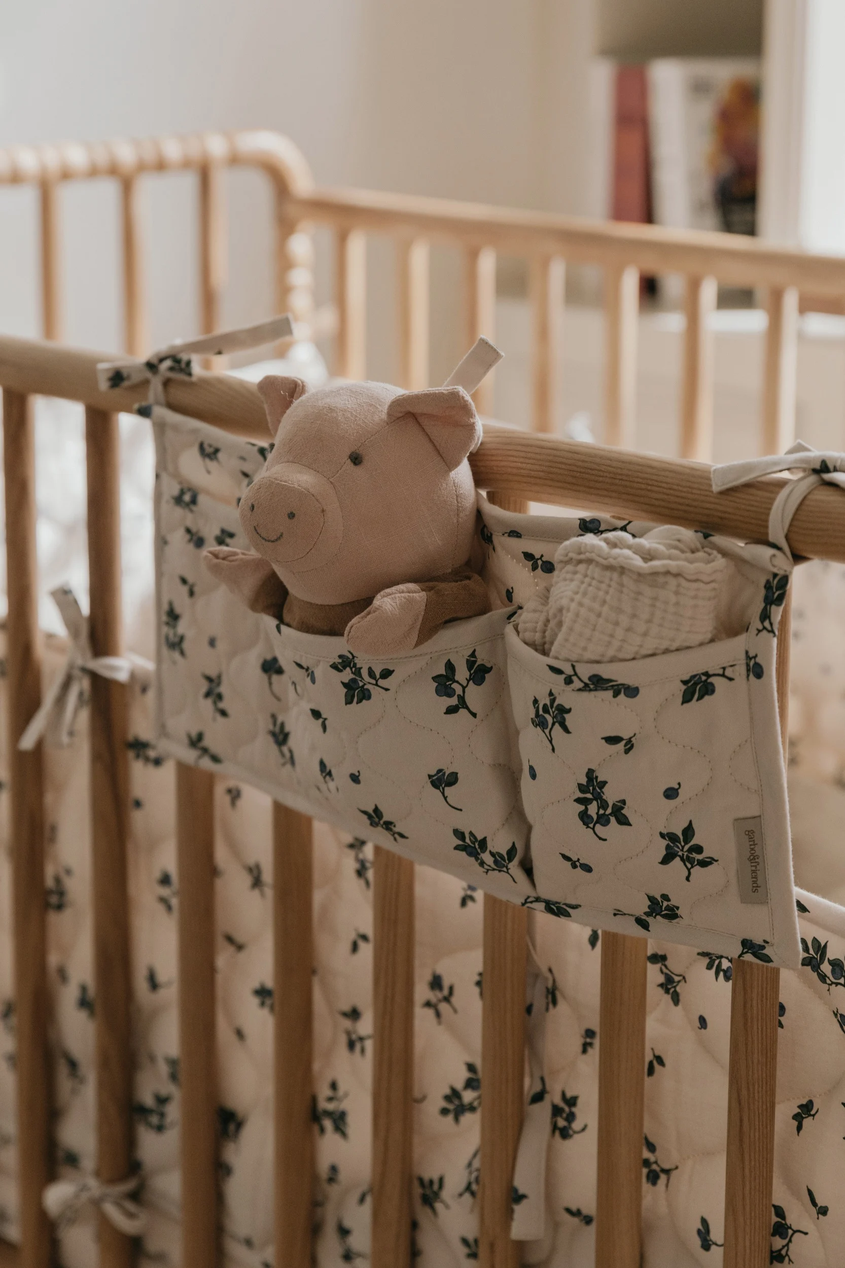 Un cerdito de peluche rosa y una tela de muselina blanca en un organizador floral crema y azul colgado de una cuna de madera.