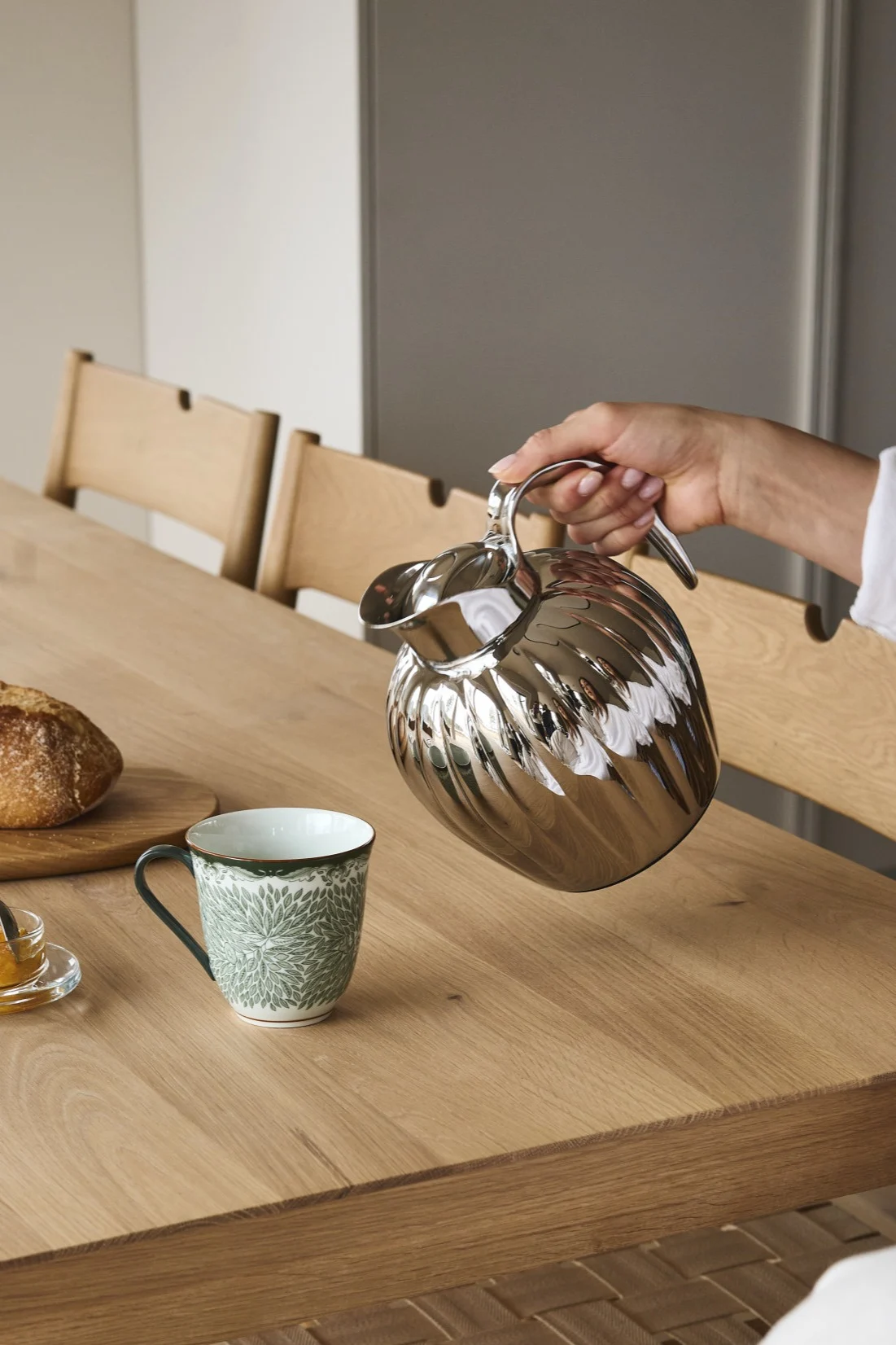 Una mano vierte de una jarra térmica plateada estriada en una taza verde estampada sobre una mesa con pan y mermelada.