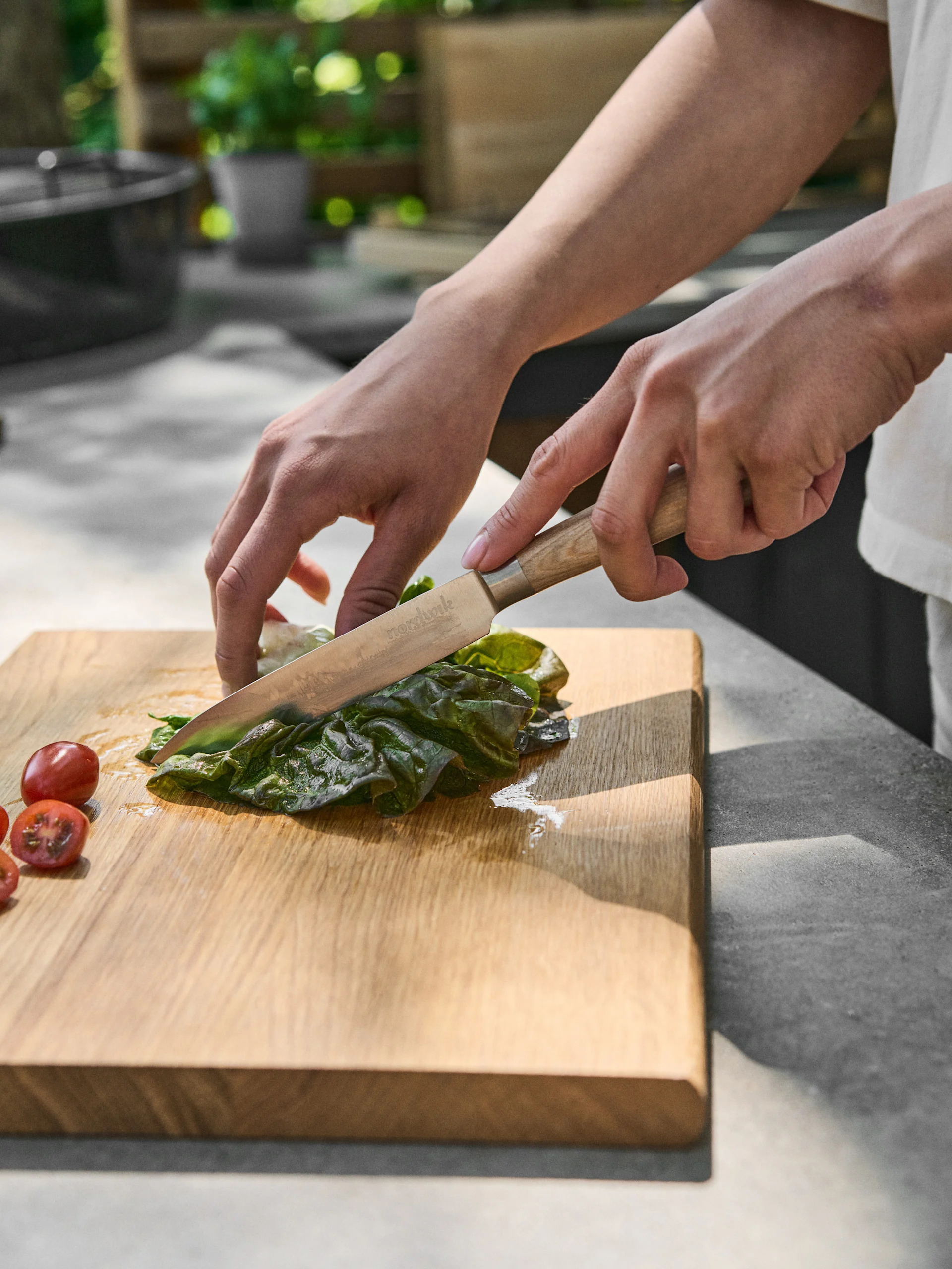 Manos cortando una hoja verde con un cuchillo en una tabla de madera, junto a tomates cherry.