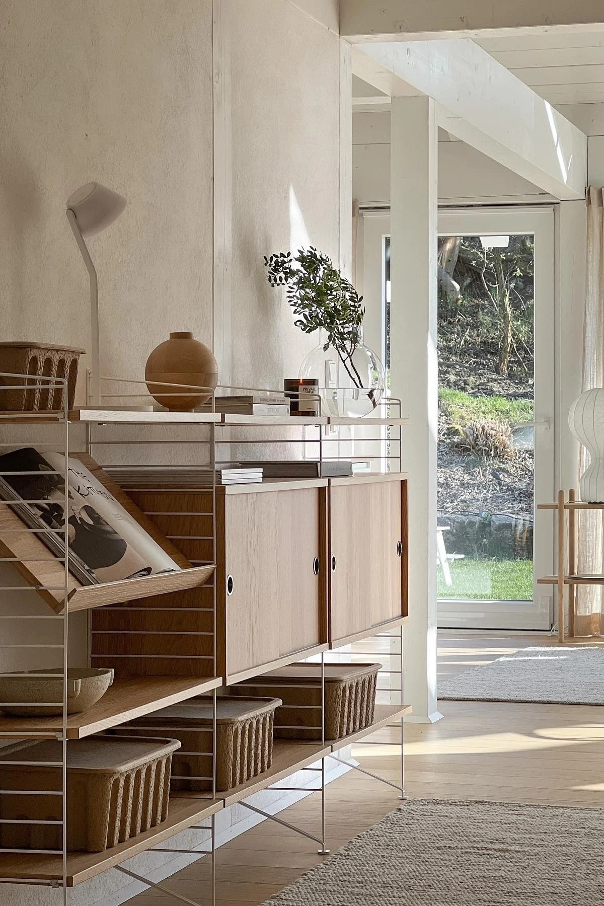 Habitación luminosa y moderna con una estantería String de madera clara y marcos blancos, llena de jarrones, libros, plantas y cestas, con vistas al jardín. La estantería es perfecta para decorar la pared. 