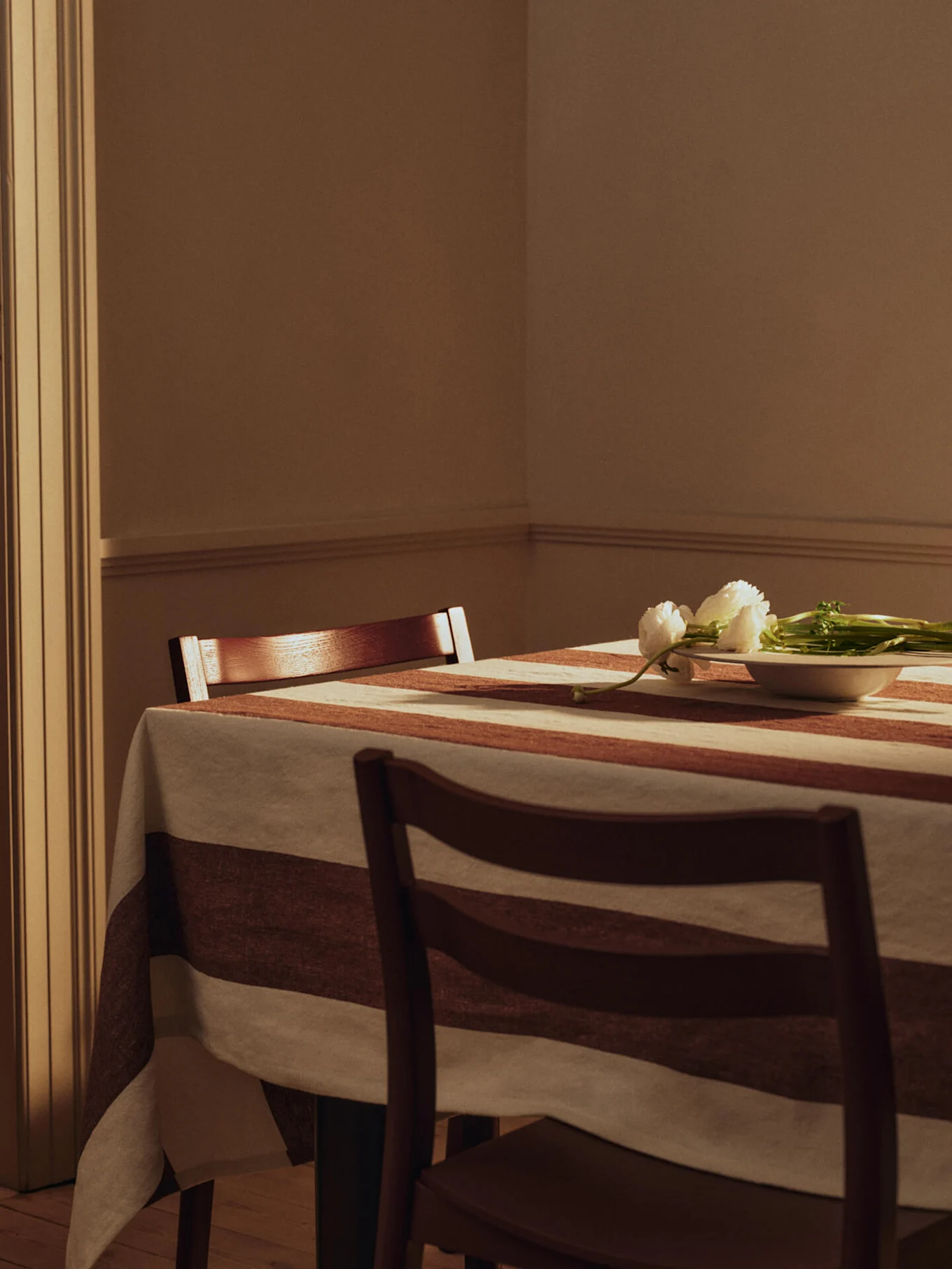 Mesa de comedor con mantel a rayas, dos sillas de madera y flores blancas en un cuenco pequeño.