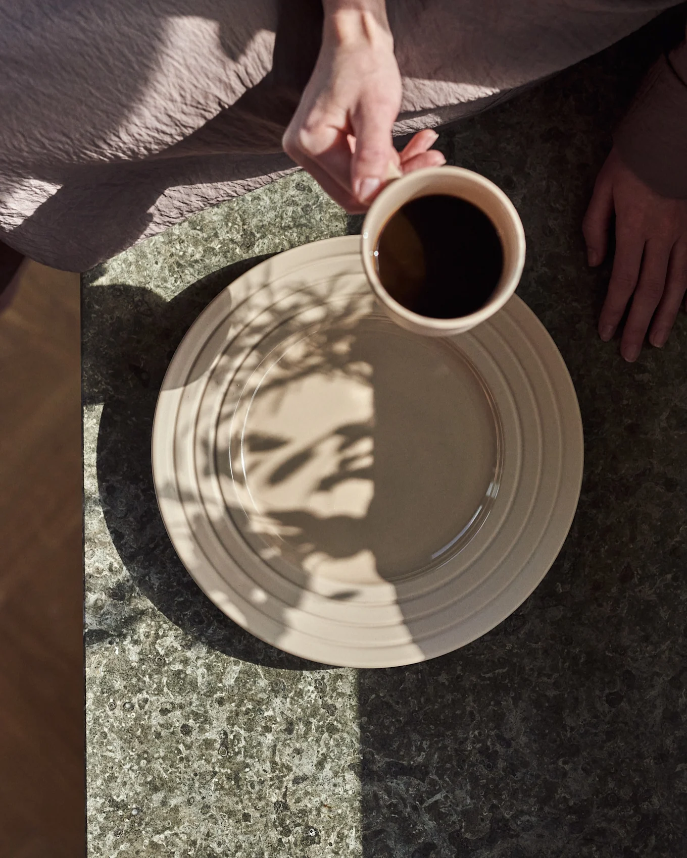 Vista cenital: manos, una sostiene una taza beige con café negro sobre un plato beige en una mesa de piedra oscura, con sombras de hojas.