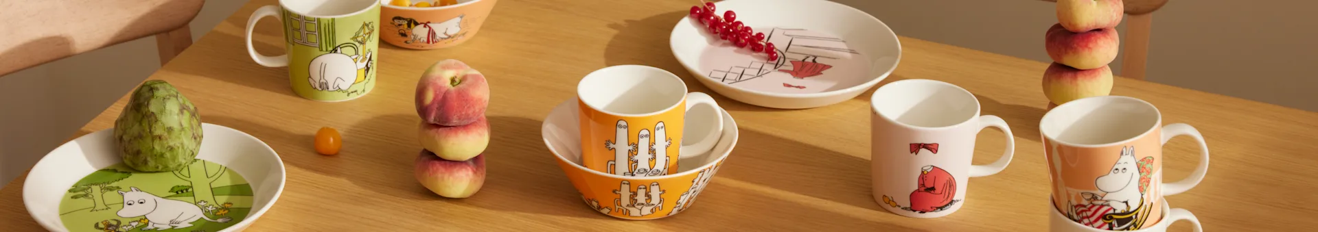 Vajilla Moomin en varios colores y diseños, con tazas, cuencos y platos, junto con fruta fresca sobre una mesa de madera.