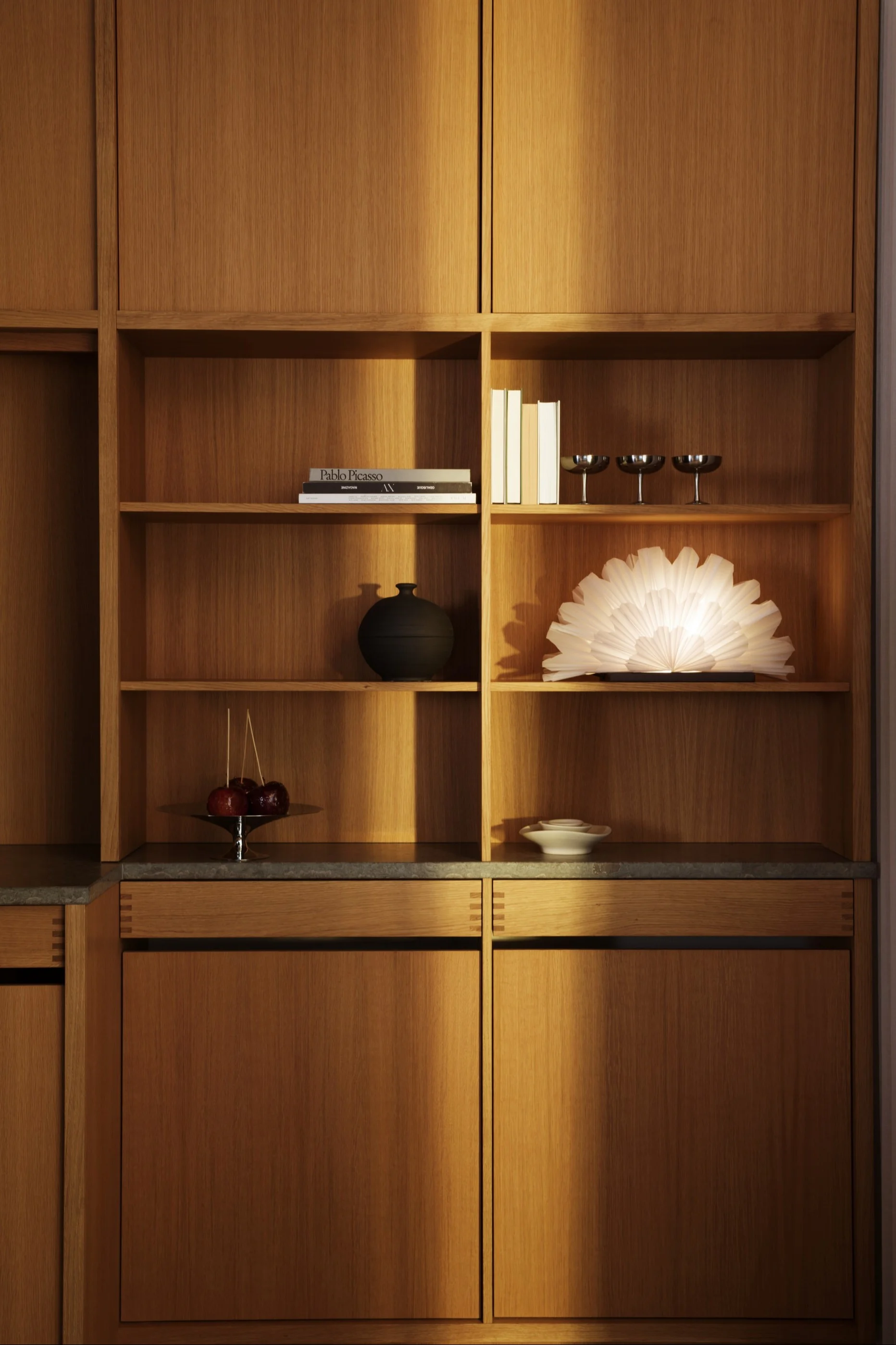Un moderno mueble de madera empotrado con estantes abiertos que exhiben libros, un jarrón negro, una lámpara brillante en forma de abanico, manzanas acarameladas y copas de plata.