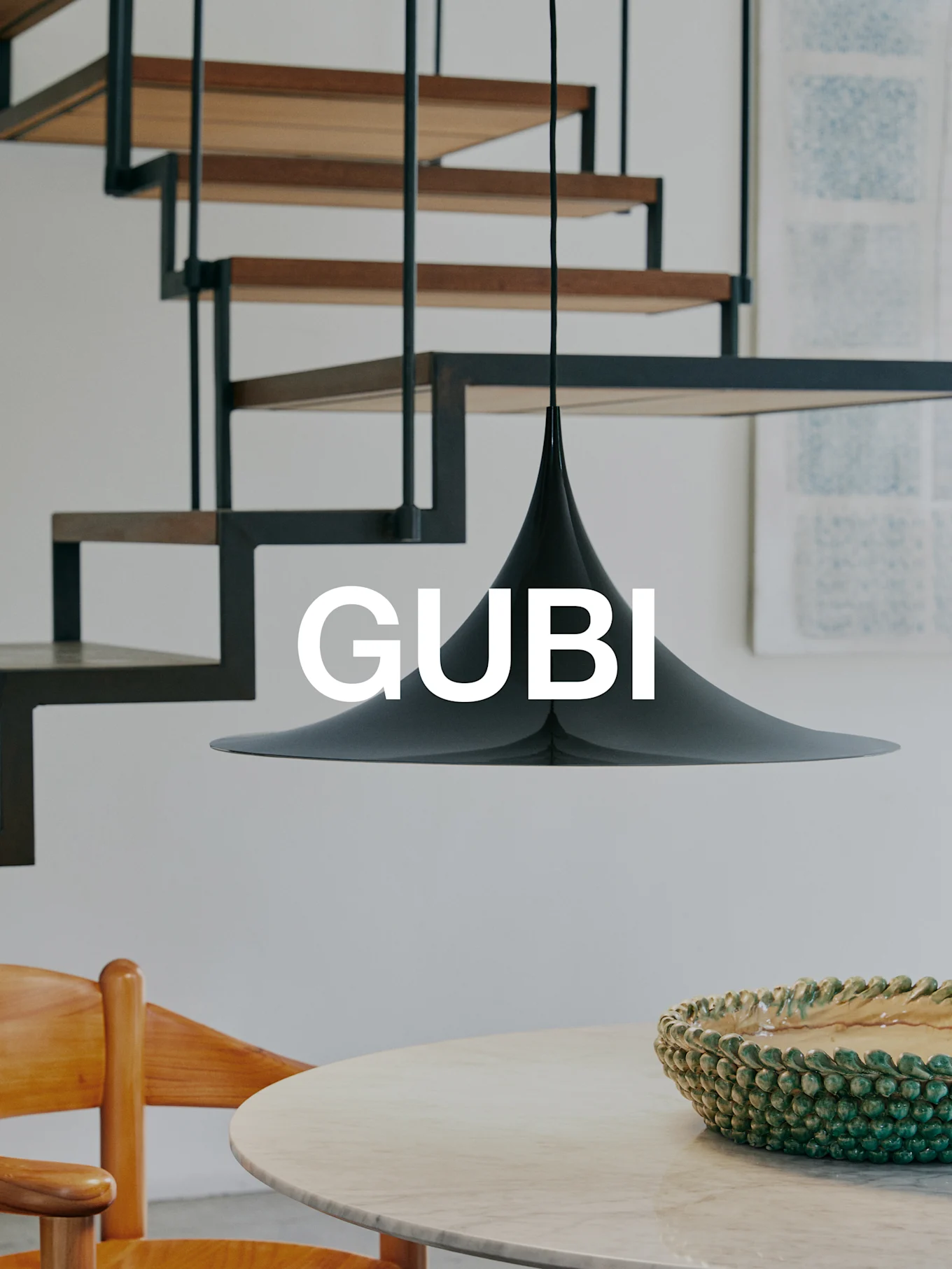 Una lámpara colgante cónica negra sobre una mesa de mármol con un cuenco verde y una silla de madera. Una escalera moderna al fondo. Texto 'GUBI'.