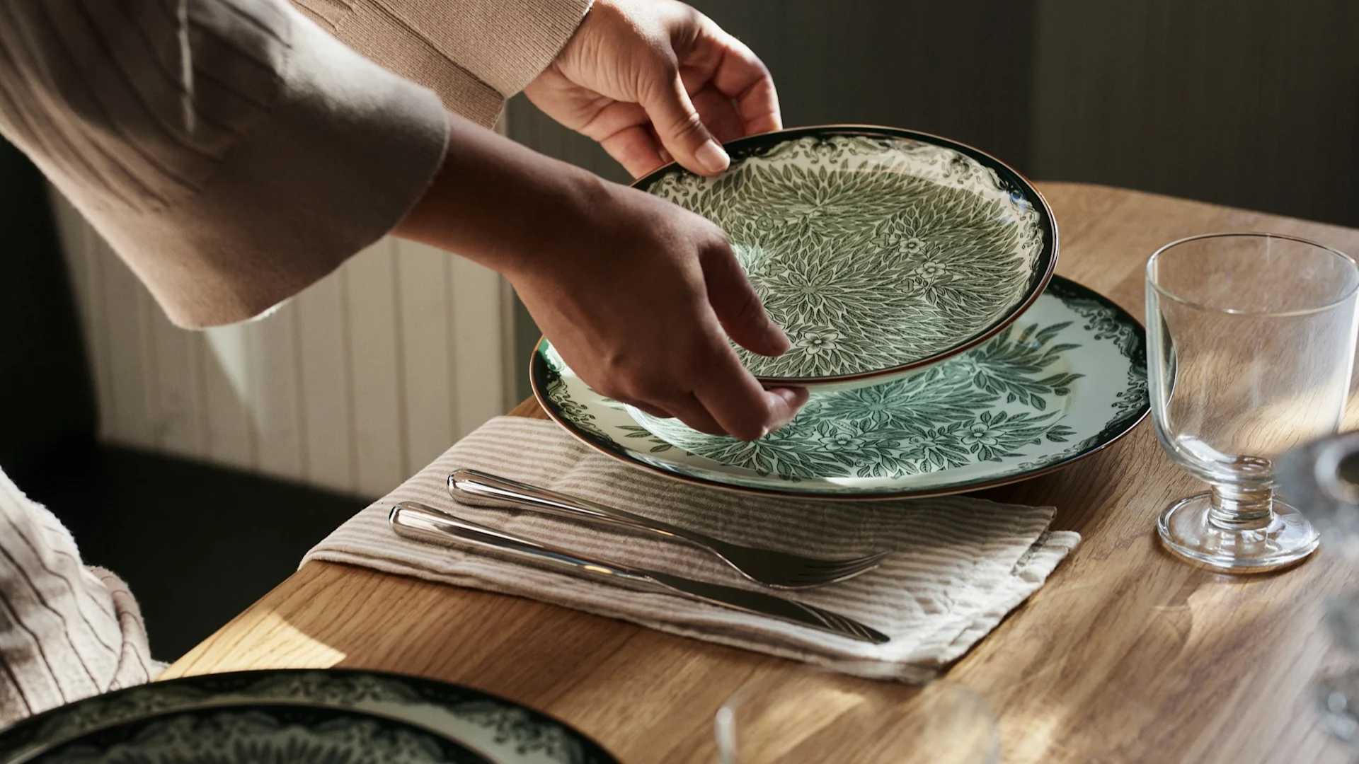 Manos colocando un plato verde y blanco sobre una mesa de madera, acompañado de cubiertos y un vaso.