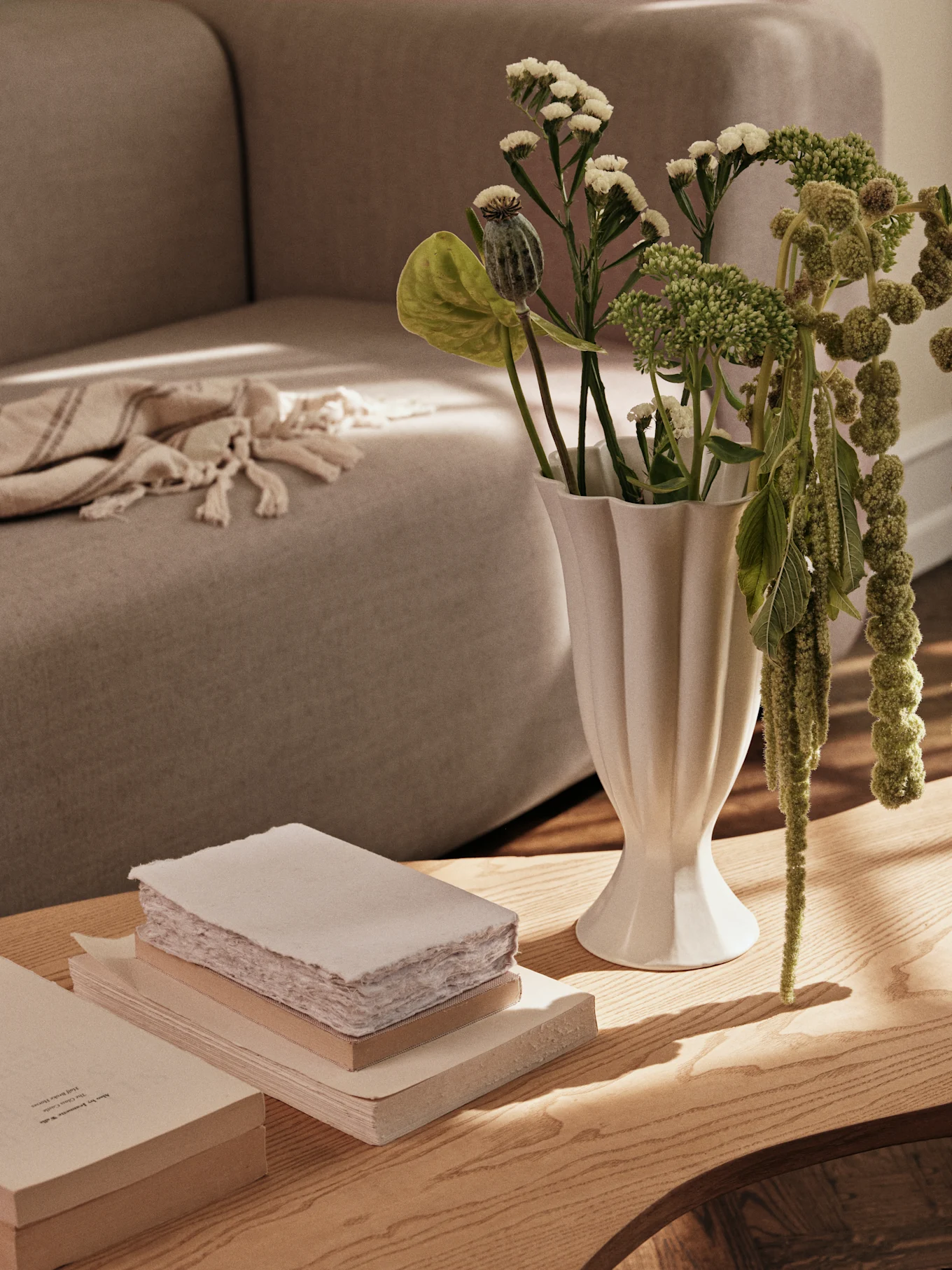 Un jarrón blanco estriado con plantas verdes y una pila de libros sobre una mesa de café de madera, con un sofá al fondo.