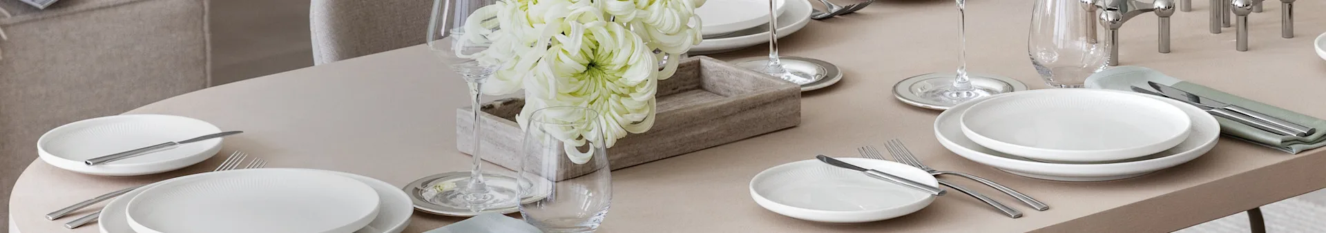 Una mesa de comedor elegantemente puesta con platos blancos, cubiertos plateados, copas de vino y vasos de agua, y un centro de mesa con grandes flores blancas en una caja de madera.