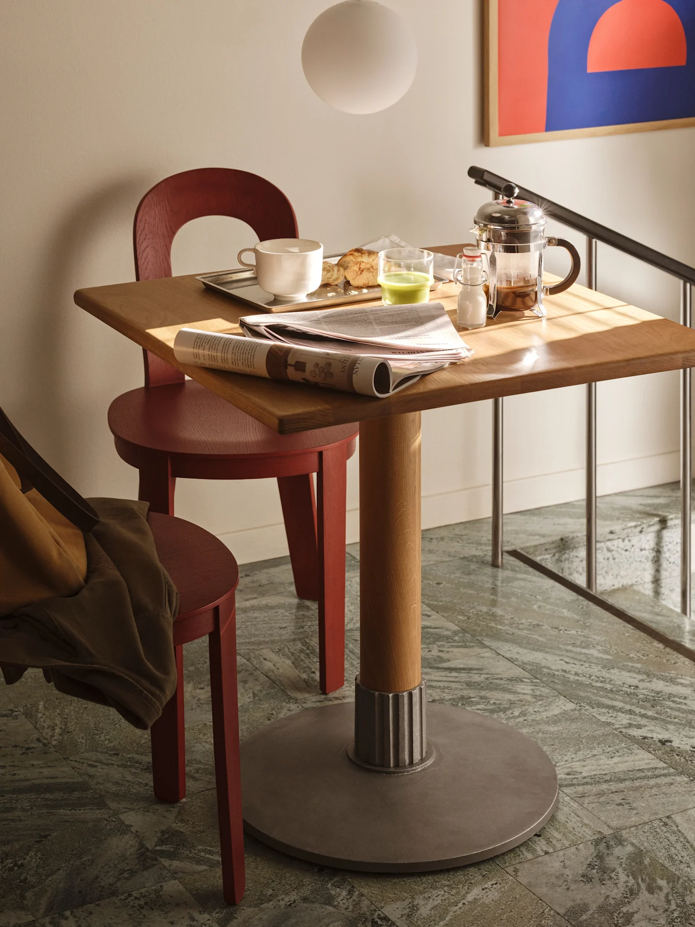 Una mesa cuadrada de madera con desayuno, un periódico y una prensa francesa, flanqueada por dos sillas rojas sobre baldosas.