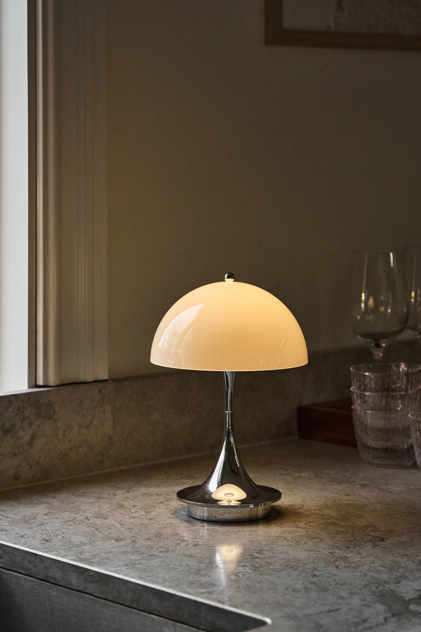 Lámpara de mesa de cromo pulido con pantalla de cúpula crema brillante sobre una encimera gris moteada, junto a una ventana y vasos borrosos.